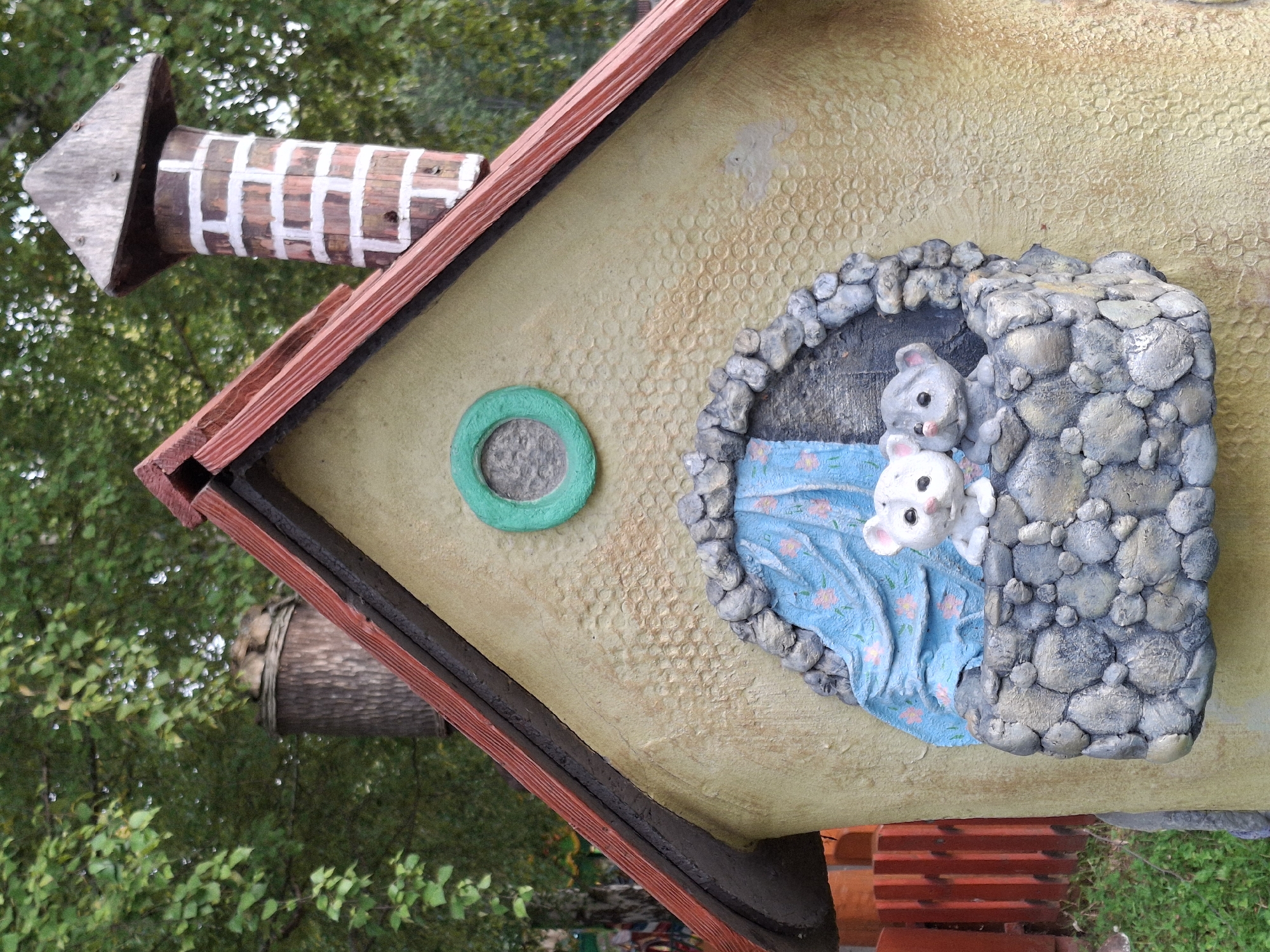 The image shows a close-up of a whimsical, cartoon-like structure, resembling a small house, with a thatched roof and a stone chimney. Two small, grey and white mouse figurines are peeking out of a window. The structure is adorned with a green circular window and what appears to be a blue curtain with pink floral patterns inside the window where the mice are situated. The exterior of the house is textured and painted a light yellow. In the background, there are green trees and a portion of a red wooden fence is visible on the left side of the frame. The overall scene suggests a playful, possibly decorative, element in an outdoor setting. This structure is located in the city of Chaikovsky, Russia.