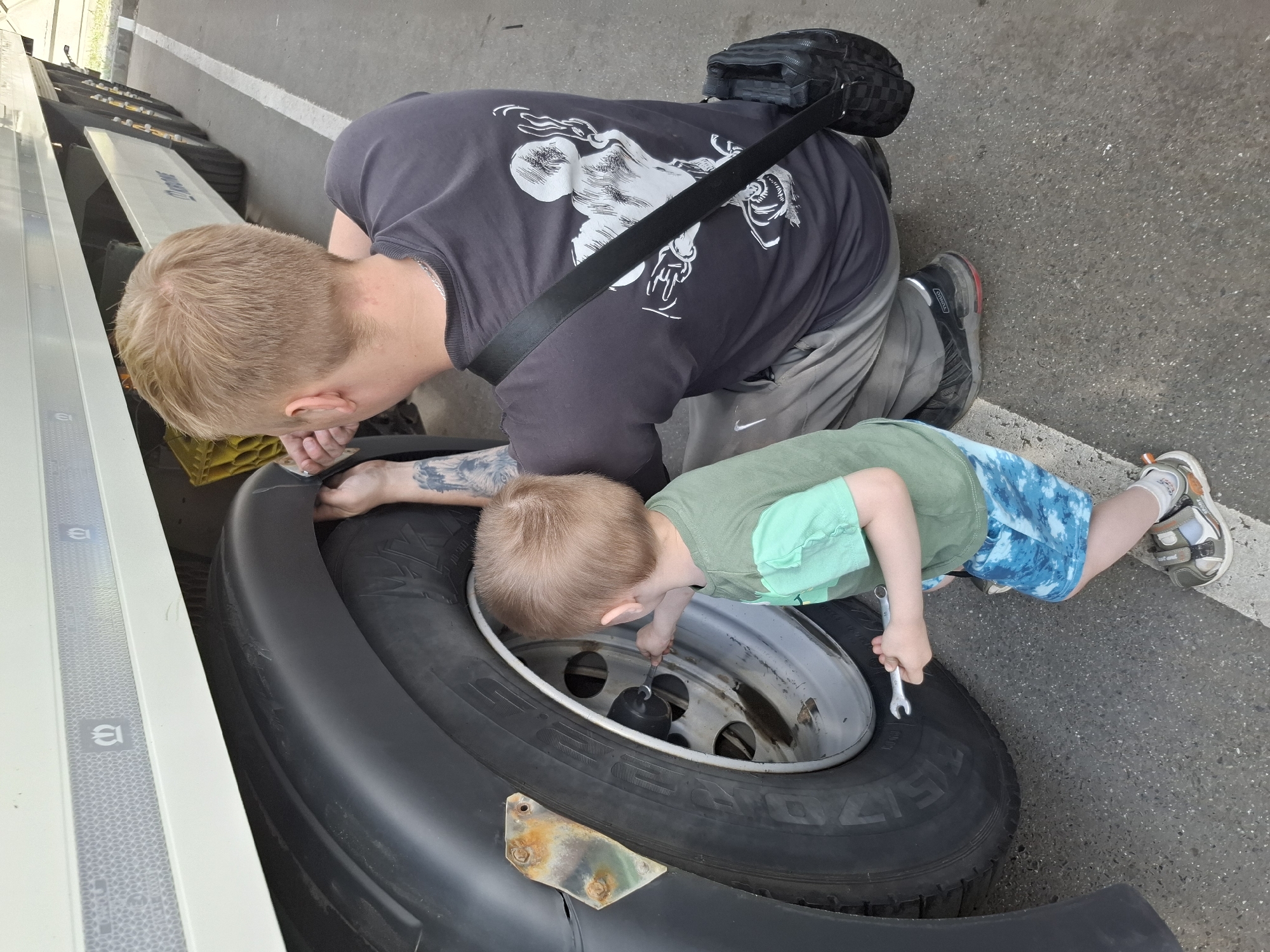 A man and a child are inspecting a tire on a large vehicle, likely a truck or trailer. The man is kneeling, wearing a dark grey t-shirt with a white design on the back, light grey pants, and black shoes with red accents. He has a black bag slung over his shoulder and a tattoo on his arm. The child is kneeling and reaching into the wheel well, holding a wrench. The child is wearing a light green t-shirt and blue shorts with a white pattern, white socks and grey and orange sandals. The tire is large and black, mounted on a silver rim. The tire is attached to a black fender. The scene appears to be outdoors, possibly a roadside or parking area, as indicated by the gray asphalt ground and a white painted line. There is a white panel of a vehicle visible to the left of the tire. There is no information in the photo to confirm it is located in Izhevsk, Russia.