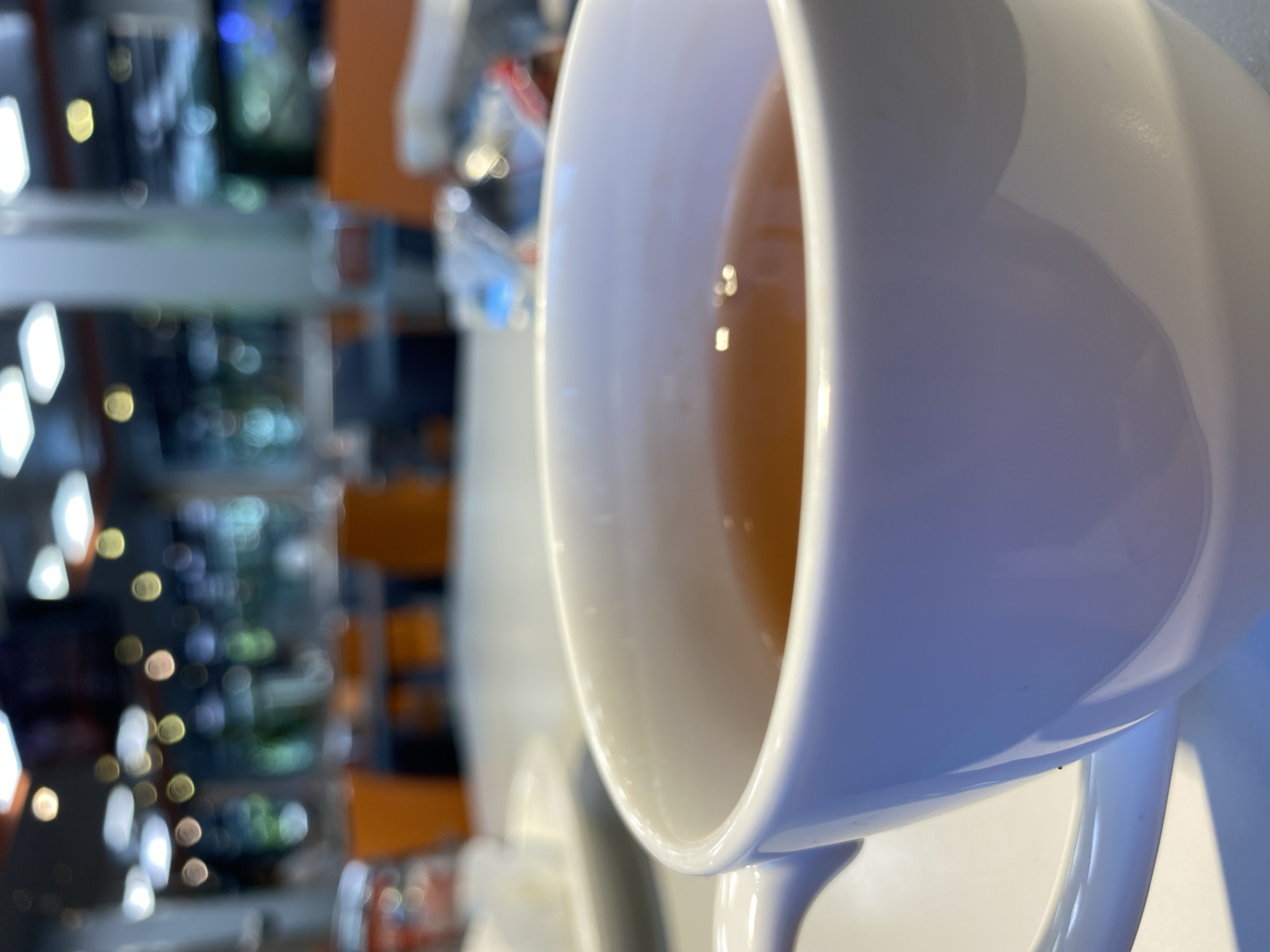 The media file shows a close-up of a white ceramic mug filled with a light brown liquid, likely tea. The mug is positioned in the foreground, with its rim and interior, containing the beverage, dominating the right side of the frame. The liquid has subtle reflections from the surrounding environment.
The background is out of focus, suggesting a shallow depth of field. It appears to be an indoor setting with shelves lined with various items, possibly in a store or a home. There are blurred lights and colors, including orange, blue, and white, which create a bokeh effect. Some of the blurred shapes hint at displays or products on the shelves, and the overall impression is of a busy or well-lit environment.
The orientation of the image is unusual, appearing rotated. No people are visible, and the focus is solely on the mug and its contents. The lighting suggests it could be daytime, but the indoor setting and the artificial lights in the background make it difficult to determine the exact time of day or weather conditions. There are no discernible location cues beyond the implied indoor setting. No text is visible in the image. The overall mood is neutral, focusing on the simple subject of a drink in a mug. The location context provided is Almaty, Kazakhstan.