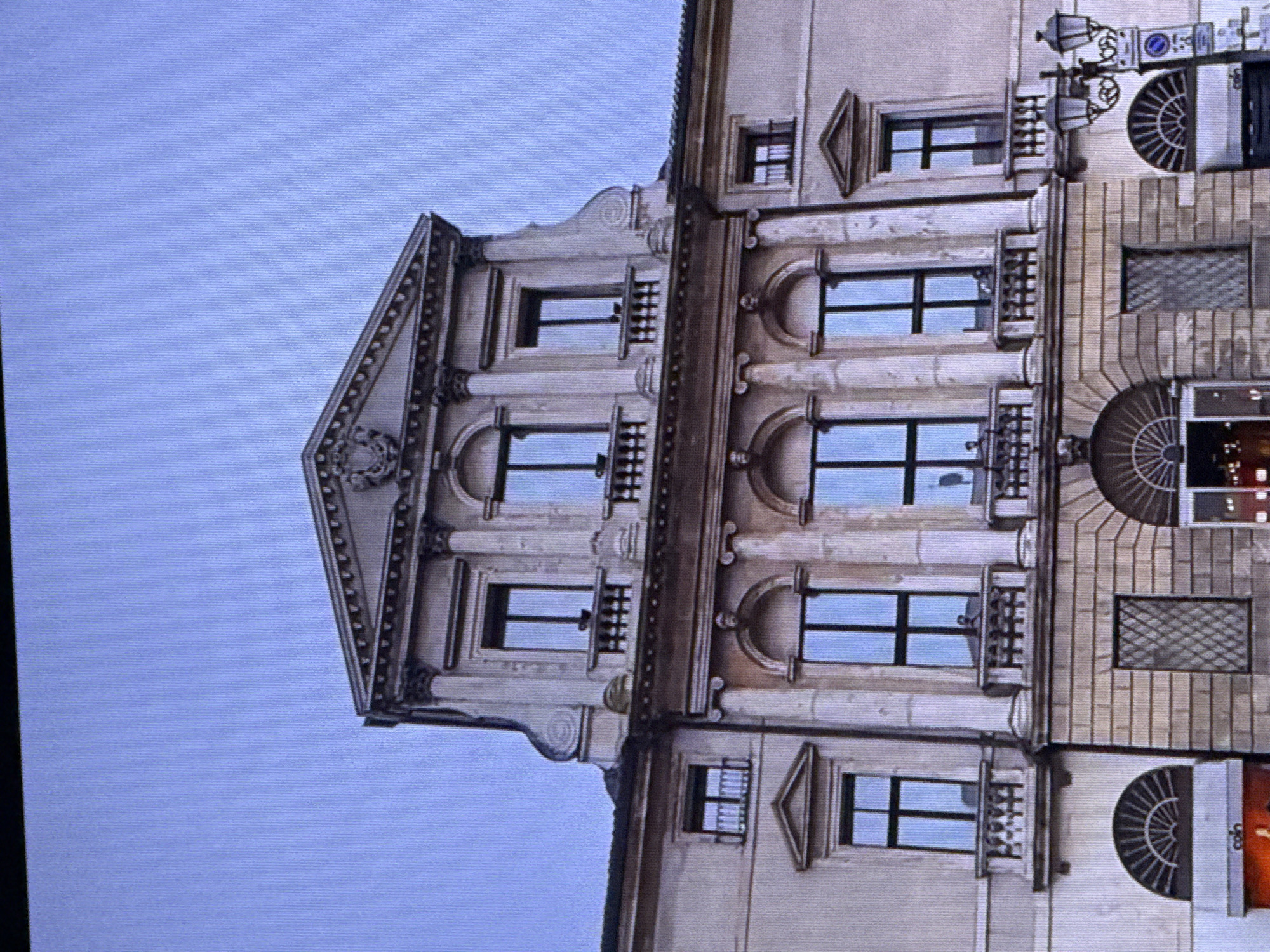 The image shows a detailed view of the facade of a building with classical architectural elements, under a clear blue sky. The building features multiple stories with ornate window frames and decorative stonework. On the upper level, there is a pediment with a coat of arms. The middle section of the building is characterized by large arched windows framed by columns. The lower part of the facade displays stone block construction with arched openings. A street lamp with multiple lights stands to the right of the building. The scene appears to be an exterior shot of a historical or grand public building. There are no discernible individuals or specific actions taking place. The location is not Dubai, United Arab Emirates, as the architecture does not align with typical Dubai styles.