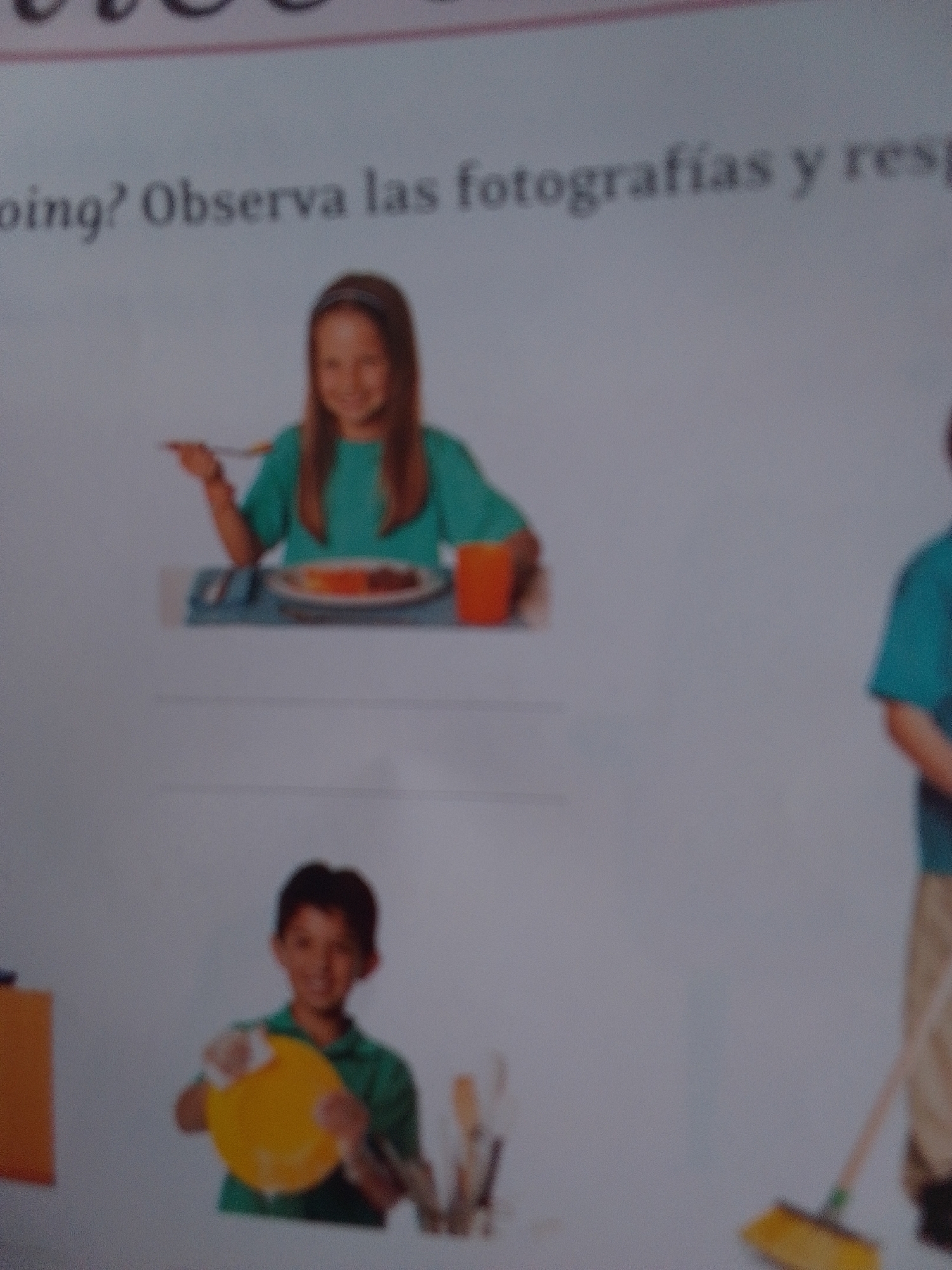 The media file displays an image with two children and a portion of a third person. The top section shows a young girl with brown hair, wearing a teal shirt and a headband, seated at a table. She is holding a fork and appears to be eating from a plate. The plate contains food, and an orange cup is visible on the right side of the table. The setting suggests a mealtime.
Below the girl, a young boy with dark hair and a green shirt is shown holding a yellow plate or bowl, with a white object, possibly a sponge or cloth, in his hand, indicating he is washing dishes.
To the right of the boy, the lower half of another person, wearing a teal shirt and dark pants, is visible, holding a broom with a yellow brush head, suggesting they are cleaning.
The image appears to be from a book or educational material, as text is visible at the top. The visible text reads: "oing? Observa las fotografías y resp". This translates to "oing? Observe the photographs and answer". The context of "La Tagua, Venezuela" suggests this is a scene from that location, possibly illustrating daily activities. The overall mood appears to be one of domestic activities, with the children engaged in eating and chores. The lighting and clarity of the image suggest it was taken indoors, possibly during the daytime.