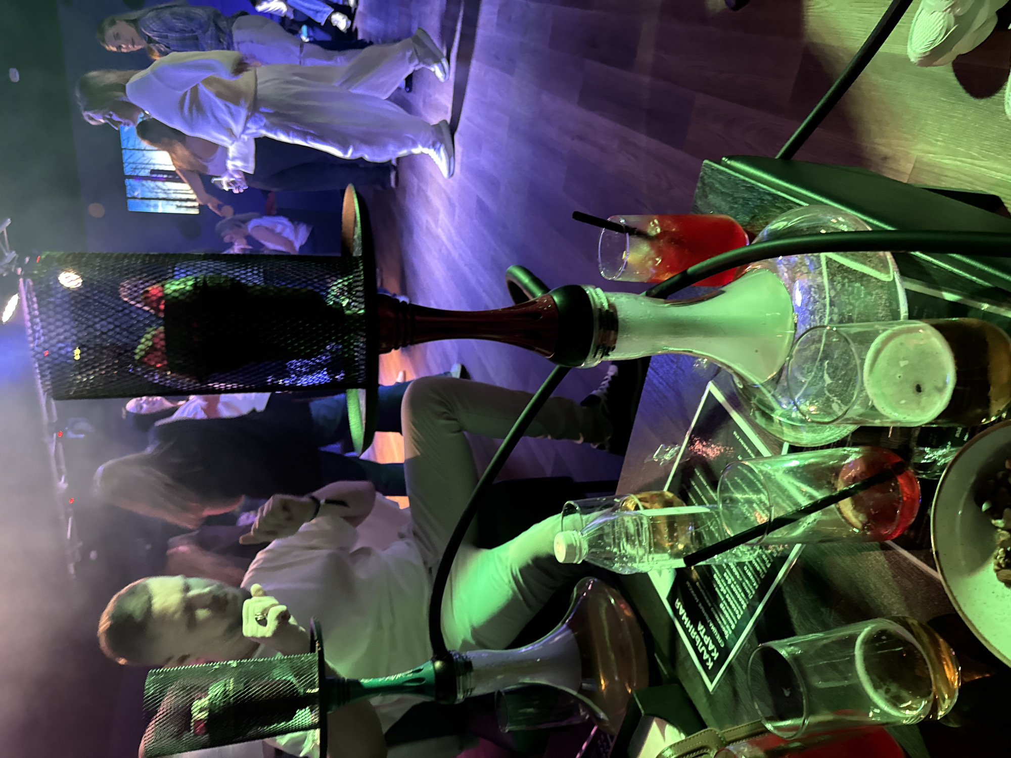 The image captures a dimly lit indoor scene, likely a lounge or nightclub in Peresvet, Russia. The main focus is on a hookah, with its distinctive coal holder and bulbous glass base, situated on a table cluttered with various objects. Scattered across the table are several empty glasses, some containing remnants of drinks, along with a small plate of snacks and a menu card written in Cyrillic script. The presence of condensation suggests the drinks were cold.
Several individuals are visible in the background, engaged in conversations or other social activities. Some are sitting while others are standing. The clothing suggests a casual yet somewhat upscale atmosphere. The interior features modern décor, and the lighting, a combination of ambient and focused beams, contributes to the relaxed and informal ambiance.