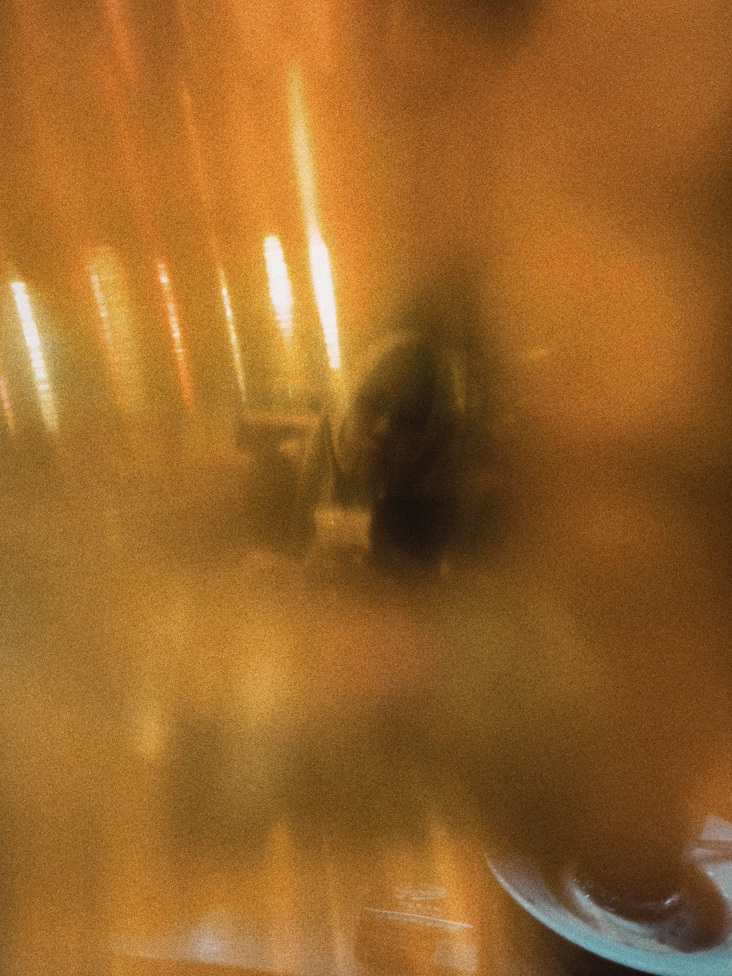 The image shows a blurry view in what appears to be a restaurant or cafe. Bright streaks of light are visible, likely from light sources within the establishment. A dark, indistinct figure sits at a table. In the foreground, a cup and saucer are partially visible, indicating the setting is likely a place where food or drinks are served. Though the exact location is difficult to ascertain with such poor clarity, the prompt specifies that the location is in the city of Lhokseumawe, Indonesia. The overall ambiance is warm due to the orange hue and lighting.
