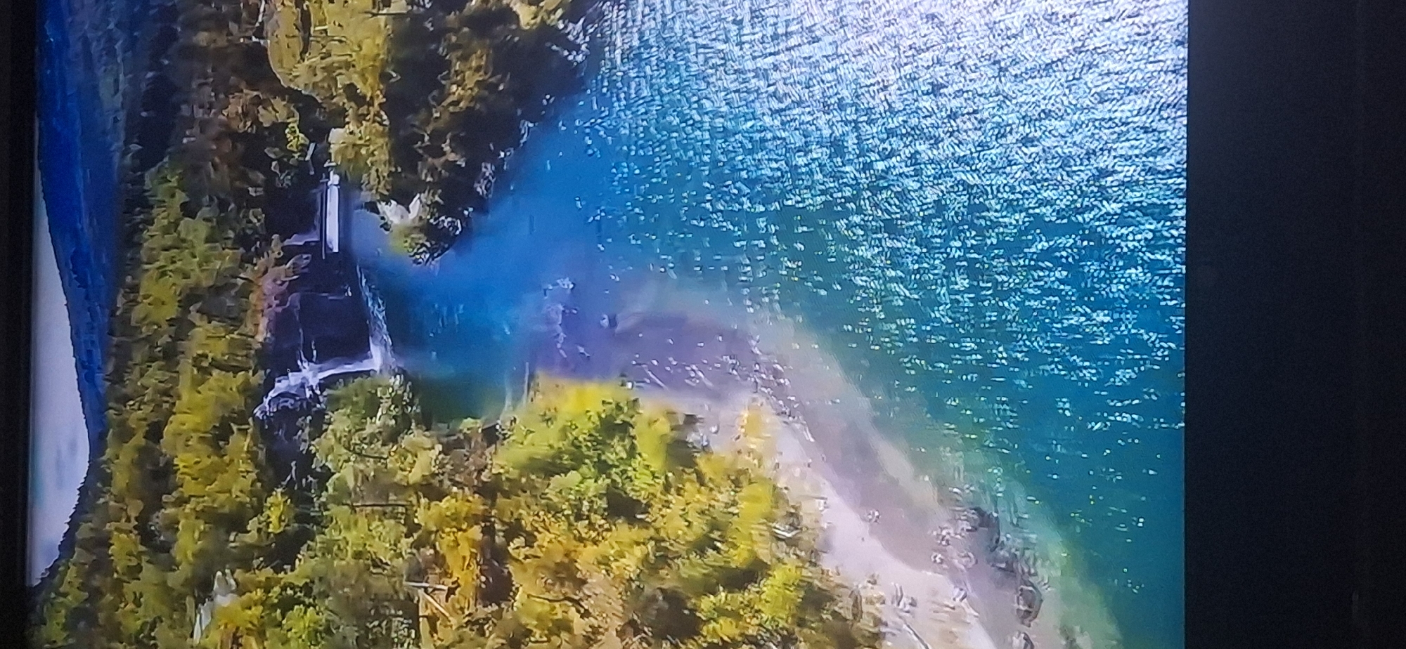This image is an aerial view of a river with a waterfall flowing into it. The river is surrounded by lush green trees and a rocky shoreline. The water is a vibrant blue, and the sunlight reflects off its surface, creating a sparkling effect. The waterfall cascades down a series of rocky ledges, creating a misty spray that is visible in the air. The scene is serene and picturesque. There is no information or context that suggests this location is in Bila Tserkva, Ukraine.