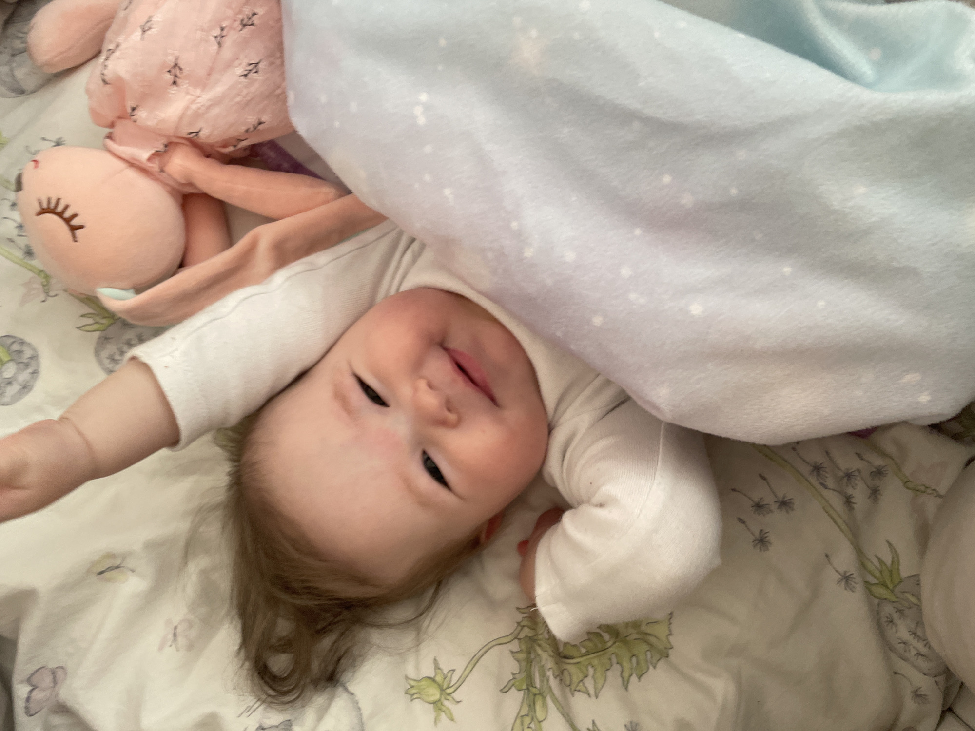 A baby in a white onesie lies on a bed with a patterned white, green, and purple sheet in an indoor setting. The baby is partially covered by a light blue blanket with white dots and a pink bunny plush toy. The location is unspecified; there is no information to indicate the location is Chaikovsky, Russia.