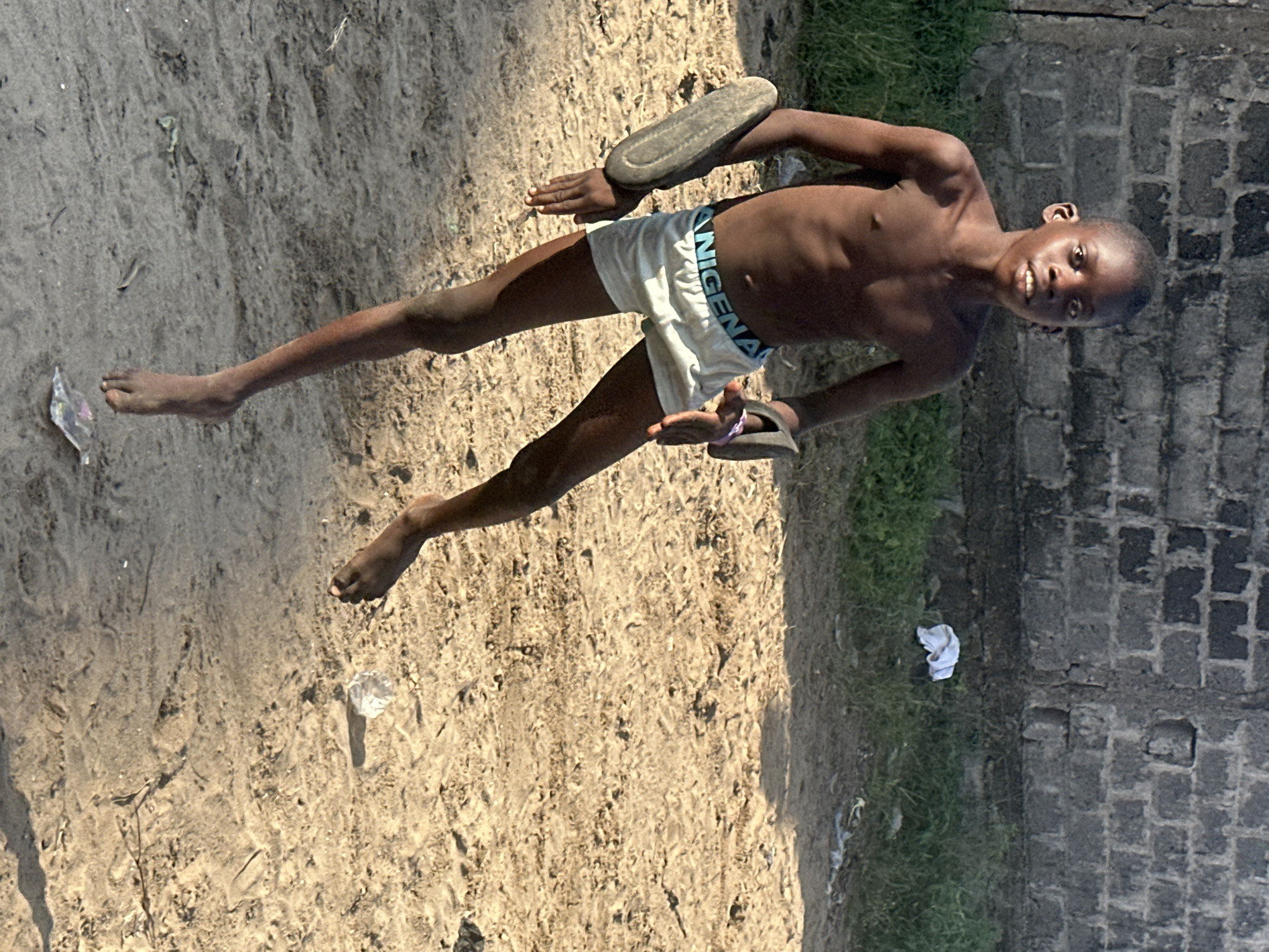 A young Black boy is shown from the waist down, wearing white shorts with "NIKE" written on the waistband. He is standing in a sandy area with some grass and a brick wall to his right. One foot is bare, while the other has a dark-colored flip-flop on it. In his right hand, he is holding another flip-flop. The boy appears to be in motion, with his legs spread apart and one arm bent at the elbow. The lighting suggests it is daytime, likely sunny, casting shadows on the sand. The background includes a grey brick wall and some patches of green grass, hinting at an outdoor setting in a possibly urban or semi-urban area of Badagry, Nigeria. There are also pieces of litter visible on the ground, including a clear plastic wrapper near his left foot and a crumpled white piece of fabric near the wall. The boy's expression, visible in a cropped image, appears to be one of playfulness or amusement as he looks towards the camera.