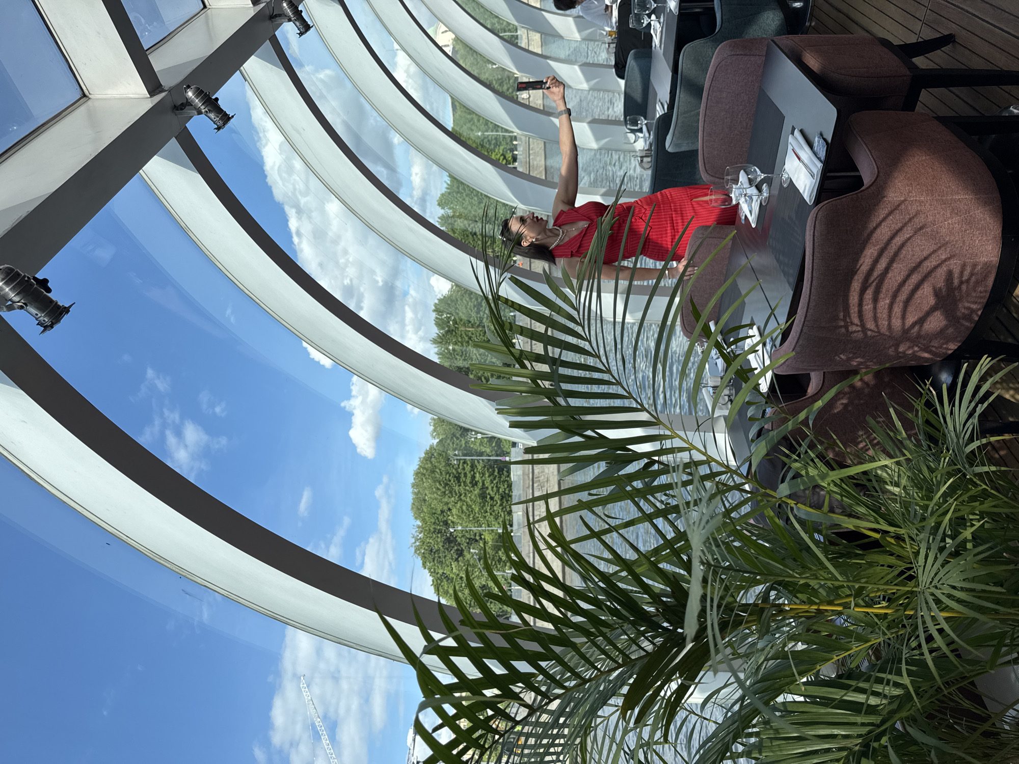 A sideways, medium shot shows a woman in a red dress and sunglasses taking a selfie with a smartphone in a building in Moscow, Russia. The building features a glass and white structural framework with reflective windows. The woman stands at a table set with glasses and napkins. Chairs surround the table. A green leafy plant occupies the foreground. Outside the windows, a body of water is visible, lined with trees. The sky is blue with clouds.