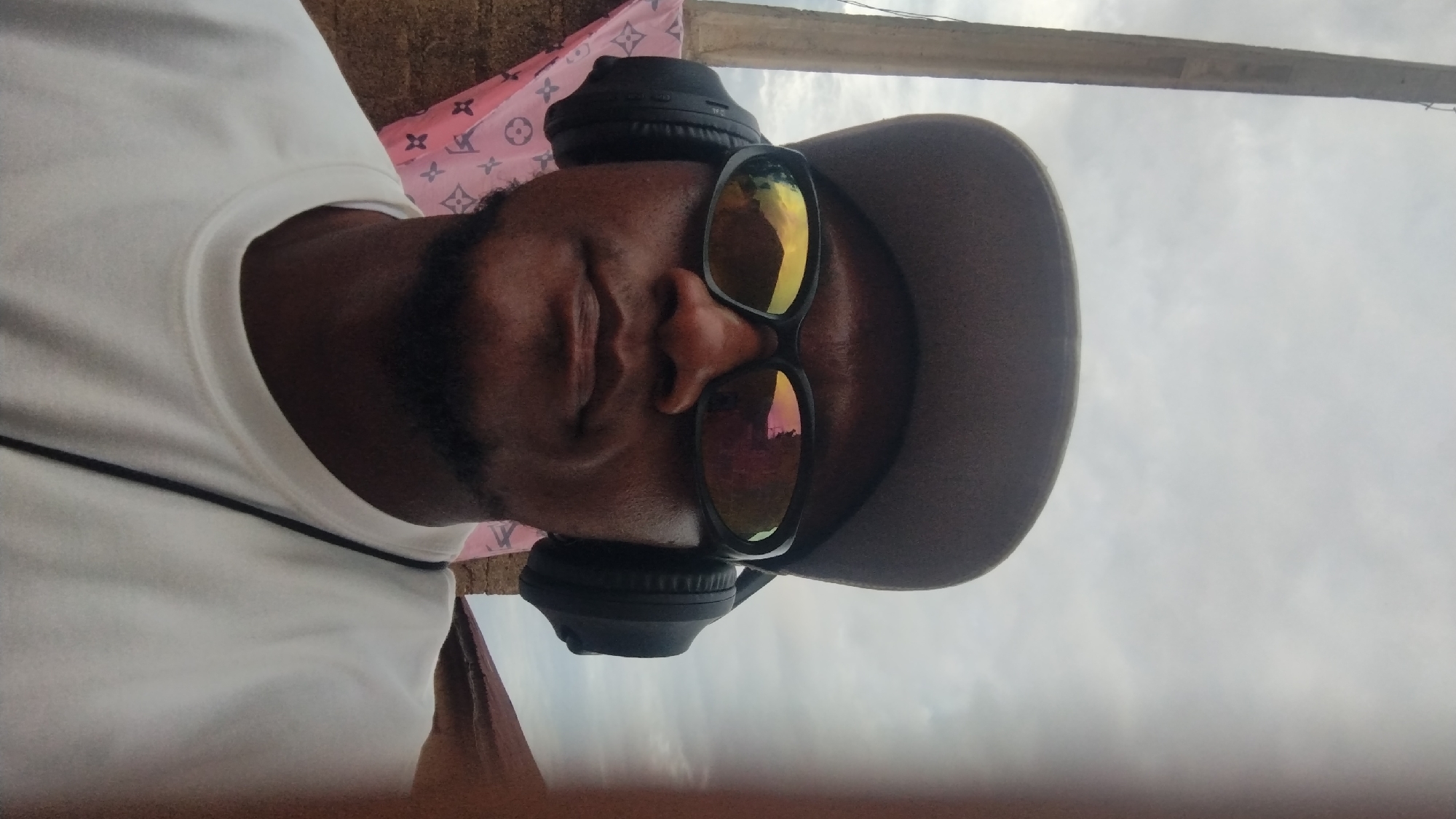 A medium, slightly angled close-up depicts a Black man wearing a grey baseball cap, black headphones, and sunglasses with reflective lenses. He has a beard. He is wearing a white t-shirt. Behind him is a pink patterned material. The background consists of a building structure with a pole and a cloudy sky. The location cannot be determined to be Jos, Nigeria based on the information given in the image.