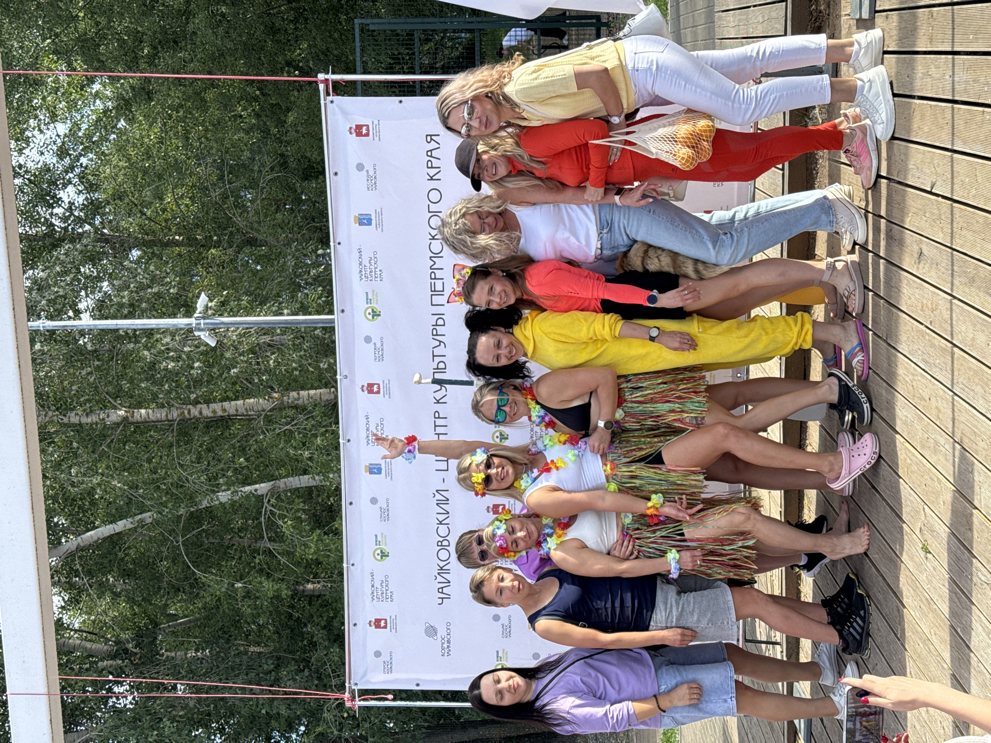 This media file depicts a group of women posing for a photo in an outdoor setting, likely during a daytime event on a sunny day. The women are arranged in a line, with some in the foreground and others behind them, suggesting a playful and informal gathering.
The subjects are all adult women, dressed in casual, summery attire. Several are wearing bright, colorful clothing, including a woman in a yellow top, another in red, and one in a bright orange top. Some are also adorned with leis and flower crowns, indicative of a festive occasion, possibly a luau or a similar themed event. One woman is wearing a grass skirt, further reinforcing this theme.
The setting appears to be outdoors, with a wooden boardwalk or deck in the background and lush green trees surrounding the area. A large banner is prominently displayed behind the women, with Russian text that includes "ЧАЙКОВСКИЙ - ЦЕНТР КУЛЬТУРЫ ПЕРМСКОГО КРАЯ" which translates to "Chaikovsky - Center of Culture of the Perm Krai." This banner provides a strong geographical context, identifying the location as Chaikovsky, Russia.
The overall mood seems cheerful and celebratory. The women are smiling and posing enthusiastically, conveying a sense of fun and camaraderie. The lighting suggests it is daytime, and the clear skies indicate good weather. The presence of the banner suggests this might be a promotional event or a gathering associated with the cultural center. The footwear visible includes sneakers and sandals, further highlighting the casual nature of the event.