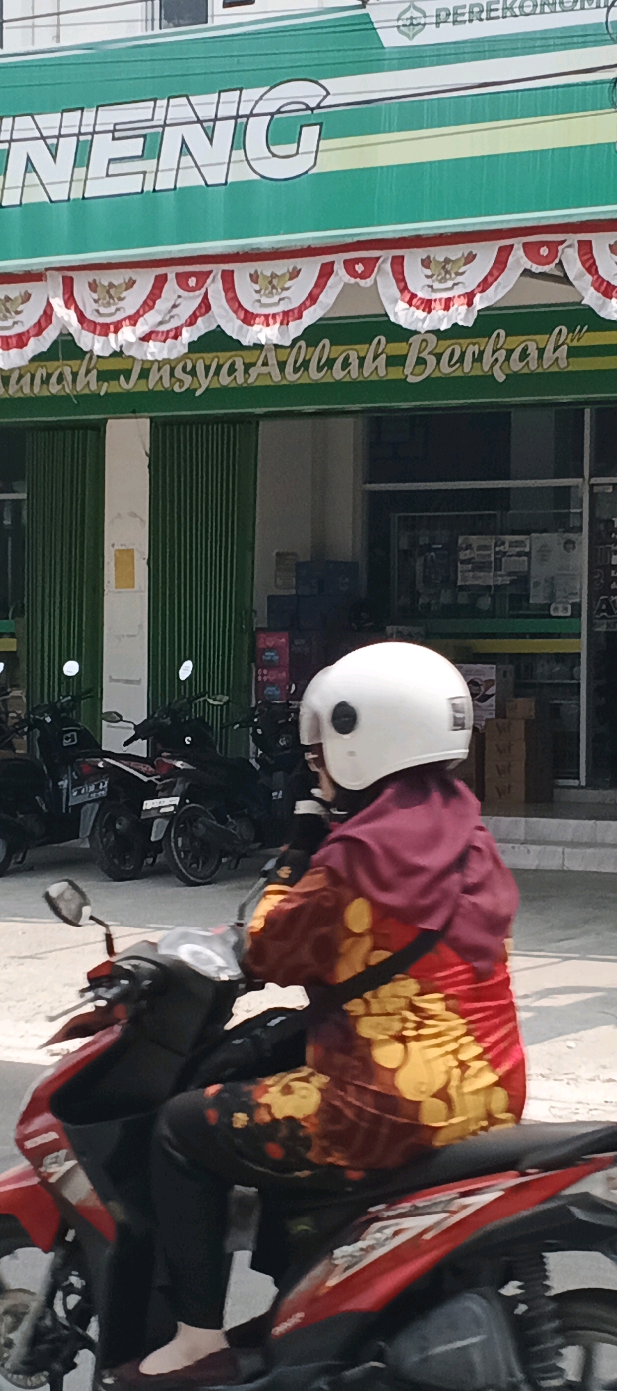 A woman wearing a white helmet and a red and yellow patterned outfit rides a red motorcycle. She is seen from the side, with her right hand on the handlebars. The woman is in front of a green building with a sign that reads "KARANGGENENG" in large white letters. Below the sign, there is a string of red and white decorative flags. The building appears to be a store or business. Several other motorcycles are parked behind the woman. The scene is outdoors, under a bright sky.