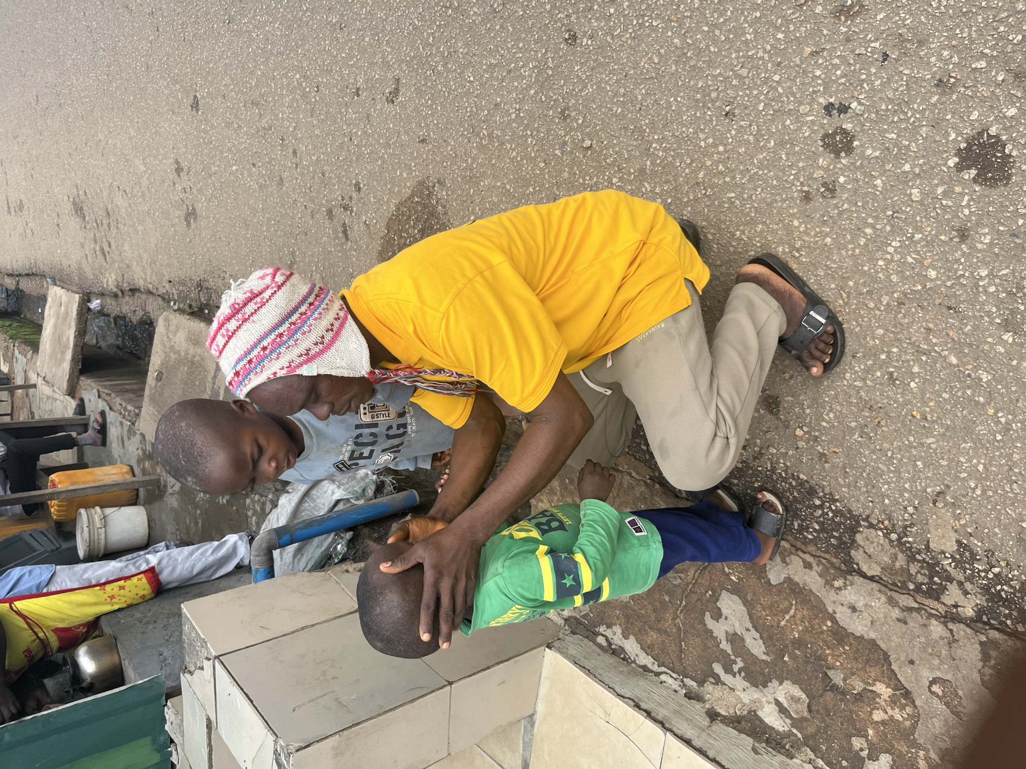 Two young Black children are the subjects of this image, which appears to be taken outdoors on a paved surface, likely a street or walkway, in Jos, Nigeria. One child, wearing a green long-sleeved shirt with yellow accents and dark blue pants, is lying on their back with their head resting on a tiled surface. The other child, wearing a light blue t-shirt with graphic print and a multi-colored knitted cap, is kneeling and cradling the head of the child lying down. A person in a bright yellow t-shirt and tan pants is kneeling behind the child in the blue t-shirt, with their hands on the child's head, appearing to be offering comfort or assistance.
The scene is somewhat cluttered, with various objects visible in the background, including buckets, containers, and what looks like folded fabric or clothing. A blue pipe is also visible near the children. The ground is a rough, grey asphalt with some darker patches and cracks. The lighting suggests it is daytime, possibly overcast, as there are no harsh shadows. The overall mood is one of care and concern, with the kneeling individuals focused on the child lying down. The graphic on the blue t-shirt includes the words "GOOD STYLE".