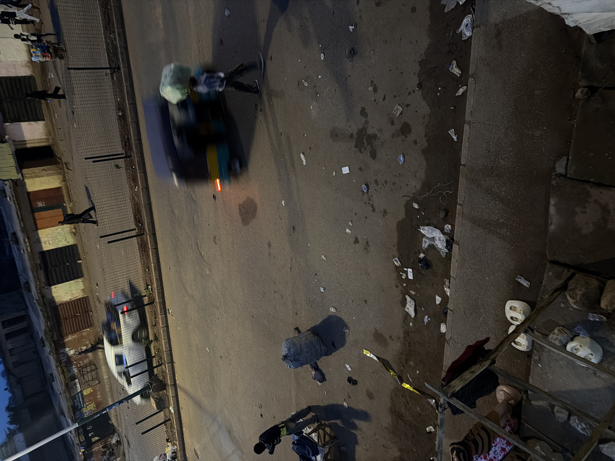 In Bukuru, Nigeria, the scene depicts a street at dusk. A white pickup truck is seen driving on the road, with its brake lights illuminated. In the foreground, a person in a blue and white checkered shirt is walking next to a moving motorized tricycle, which is carrying a large, green, plastic-wrapped bundle on its roof. Further down the road, a person in dark clothing is walking away from the camera. On the sidewalk to the left, a person in dark clothing with a yellow safety vest is standing near a cart. A large, grey, cylindrical object, possibly a sack of goods, is lying on the ground next to the person. Debris, including plastic bags and wrappers, is scattered across the road and the edge of the sidewalk. The background shows buildings with storefronts, some with corrugated metal doors.