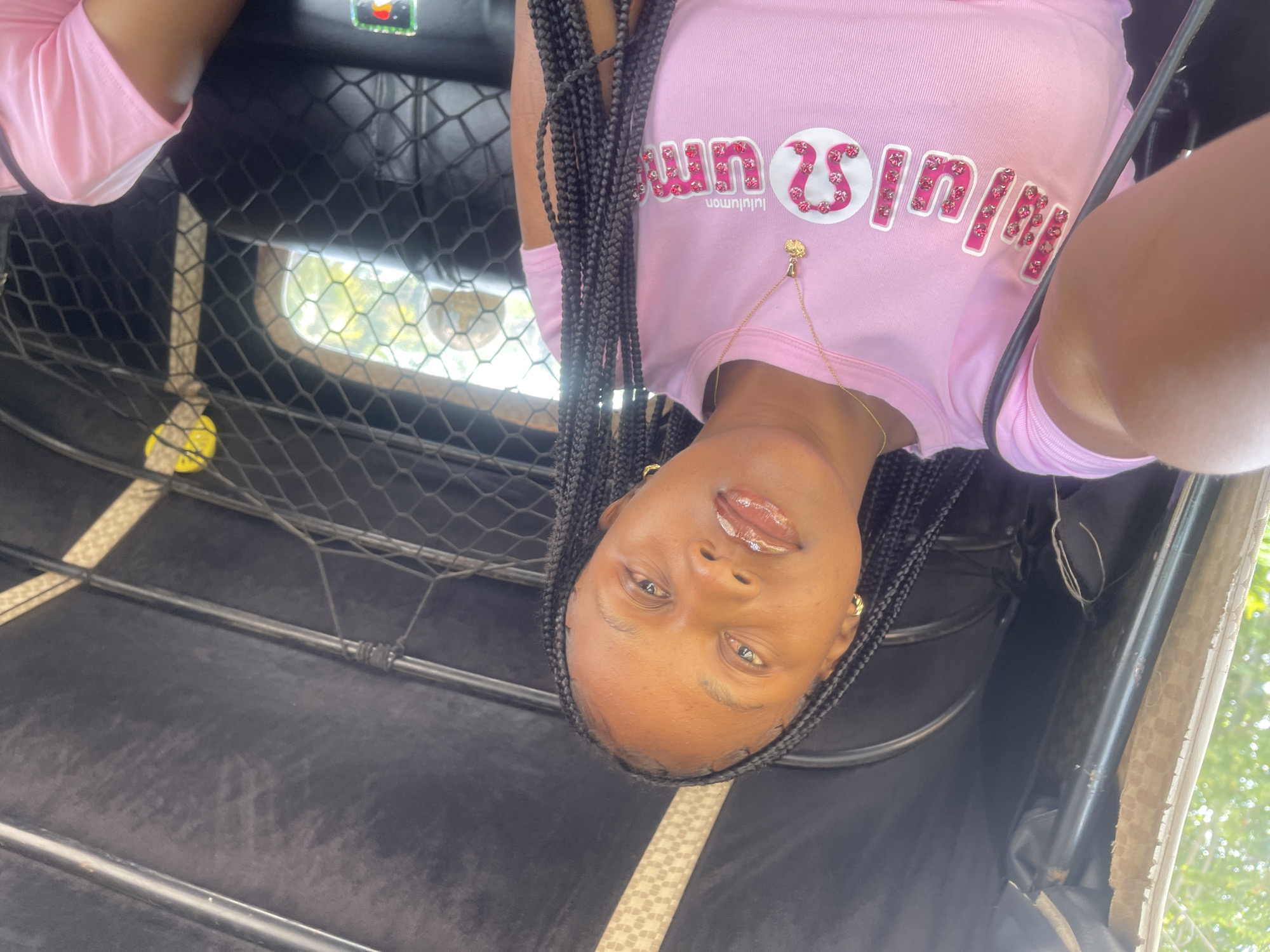 In Barakin Lerawi, Nigeria, a young Black woman with braided hair and gold hoop earrings is captured in a selfie from inside a motorized rickshaw. She is wearing a pink t-shirt adorned with the "Lululemon" logo, embellished with pink rhinestones, and a delicate gold necklace with a small pendant. Her lips are glossy, and she gazes directly at the camera. The rickshaw's interior is visible, with black upholstery and a safety net. Sunlight filters in from the background, illuminating the scene.