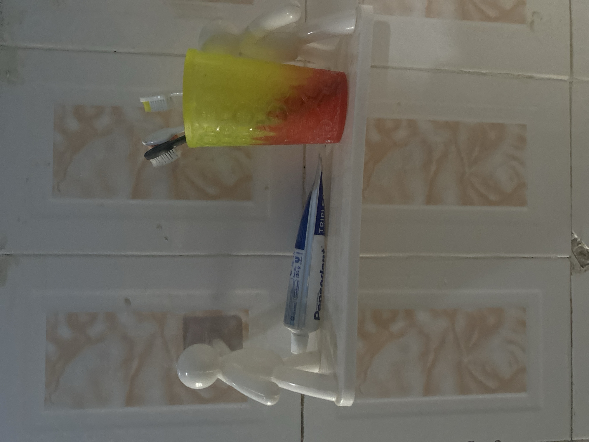 The image shows a white shelf mounted on a tiled wall. On the shelf, there is a tube of Pepsodent toothpaste, a yellow and red gradient cup with toothbrushes, and a white figurine shaped like a person. Another white figurine is partially visible to the right of the cup. The wall tiles are light-colored with a subtle marble pattern. The lighting is dim, suggesting an indoor setting, likely a bathroom. The scene is static and appears to be a simple arrangement of toiletries. There is no information in the image to suggest a location in Jos, Nigeria.