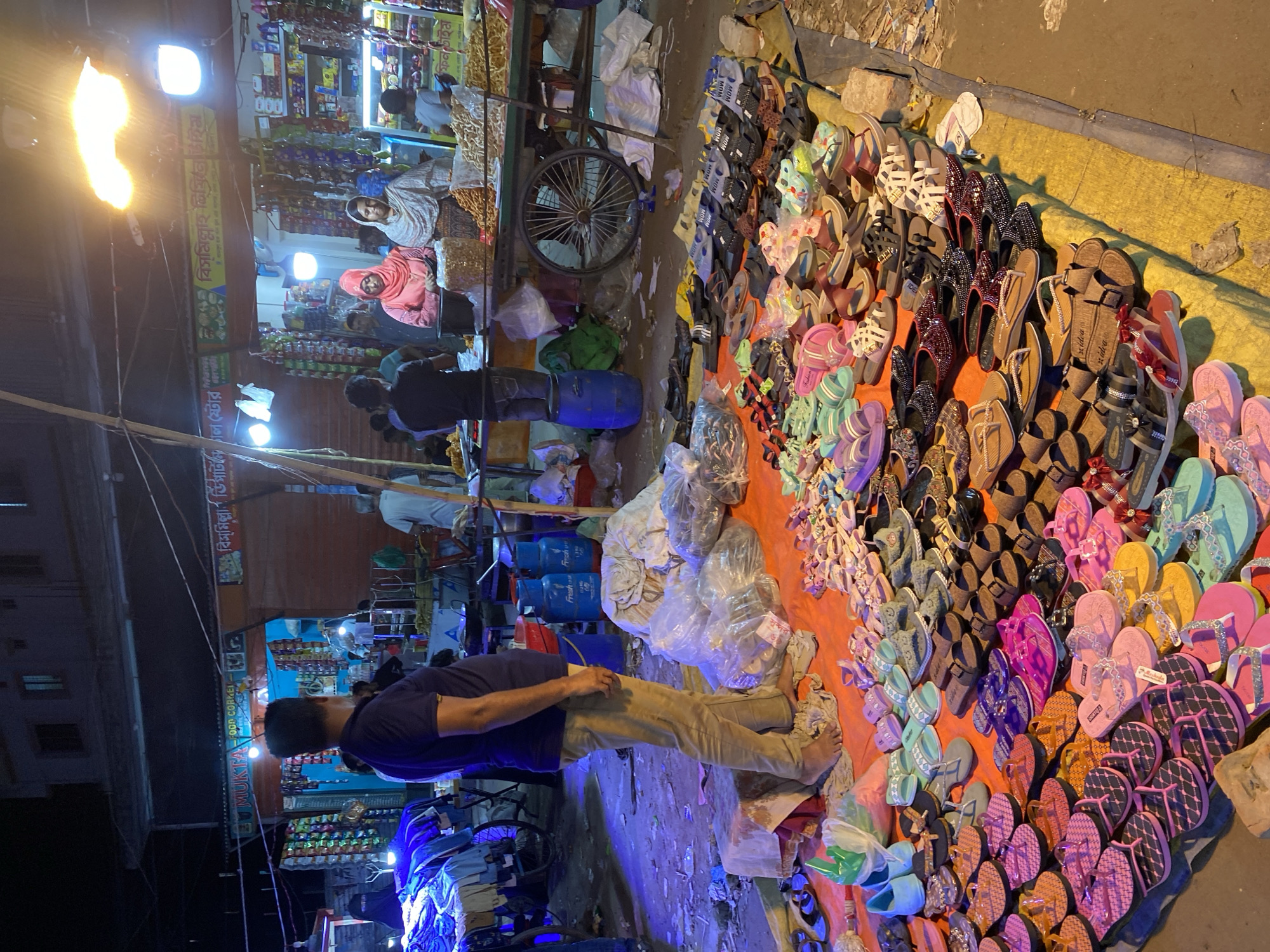 This media file captures a vibrant street market scene at night, likely in Dhaka, Bangladesh, given the provided context. The image is dominated by a large display of footwear on the right side, laid out on an orange tarp directly on the ground. This display features a wide variety of colorful sandals and flip-flops, including pink, blue, yellow, black, brown, and white designs, many adorned with glitter, bows, or intricate straps. Brand names like "WOW" and "idea" are visible on some of the shoe soles and straps.
In the mid-ground, a man wearing a dark blue t-shirt and khaki pants stands beside the shoe display. Behind him and to the left, the market continues with several open-front stalls and shops. One stall features a cart with a large spoked wheel and appears to be selling packaged goods, possibly snacks, displayed in clear containers. Another stall further back shows hanging items. Two women are visible behind one of the stalls, one wearing a light-colored head covering and another a pink head covering, suggesting they might be vendors or customers.
The entire scene is illuminated by artificial lights, including bare bulbs and fluorescent lamps hanging from makeshift structures, casting a warm glow and creating strong highlights, indicative of nighttime activity. Blue barrels and plastic bags filled with goods are also scattered around.
The market appears busy, with blurred figures in the background suggesting people are walking and browsing. The primary activity depicted is the selling and buying of footwear and other market goods.
Visible text on shop signs and products includes: "EV MUKTA," "FOOD CORVET" (likely "CORNER"), "বিসমিল্লা ডিপার্টনে এল স্টোর" (Bismillah Departmental Store), "বিসমিল্লাহ নিমিনে টাইম," and "Fresh Freshw" (likely "Fresh Water"). The Bengali script further confirms the regional setting.