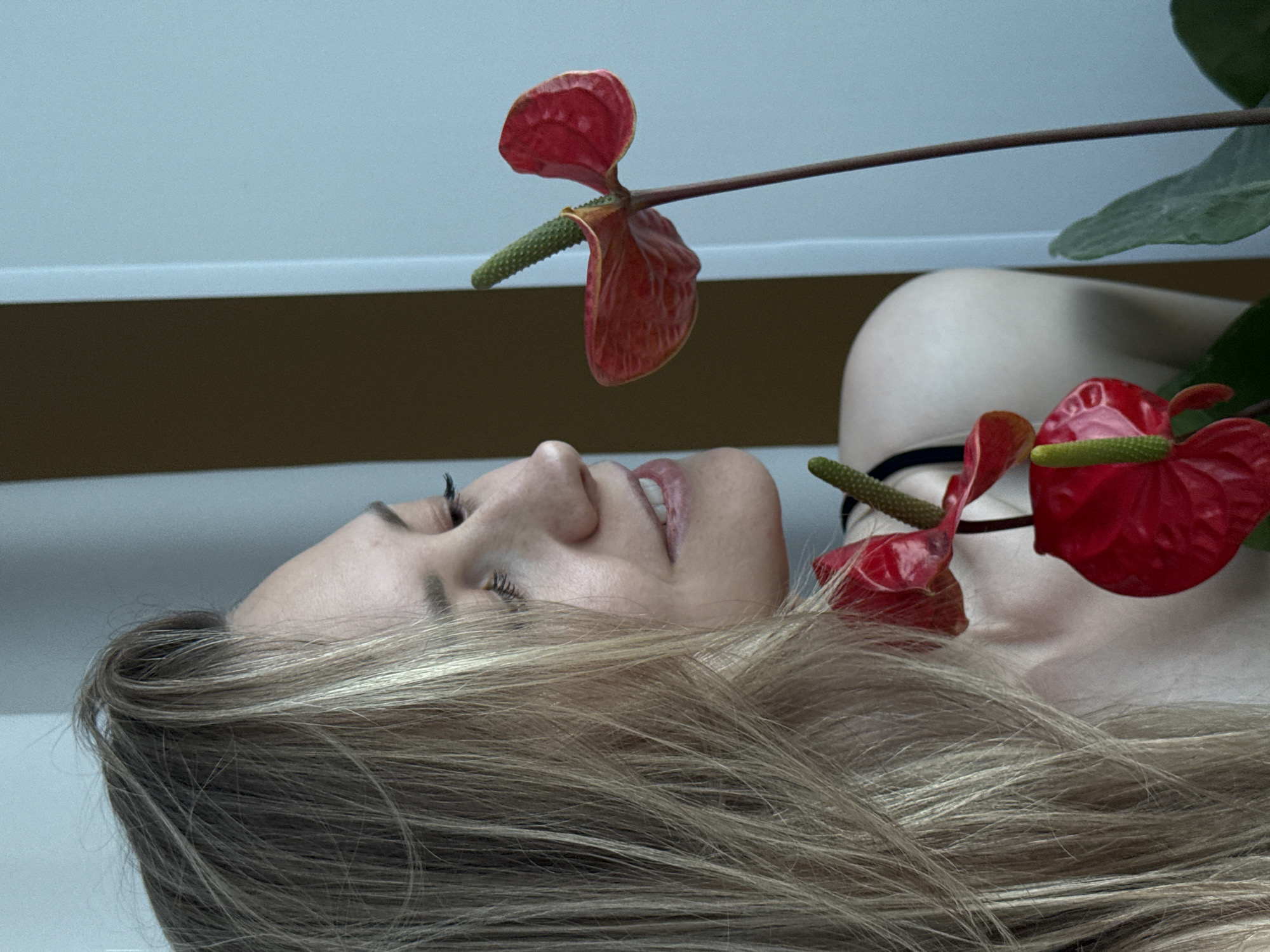 A woman with blonde hair lies on her side, facing left. Her eyes are closed and her lips are slightly parted. She is surrounded by Anthurium flowers with red spathes and green spadices. The background appears to be a light blue-gray wall with a horizontal brown accent line. The location is Chaikovsky, Russia.