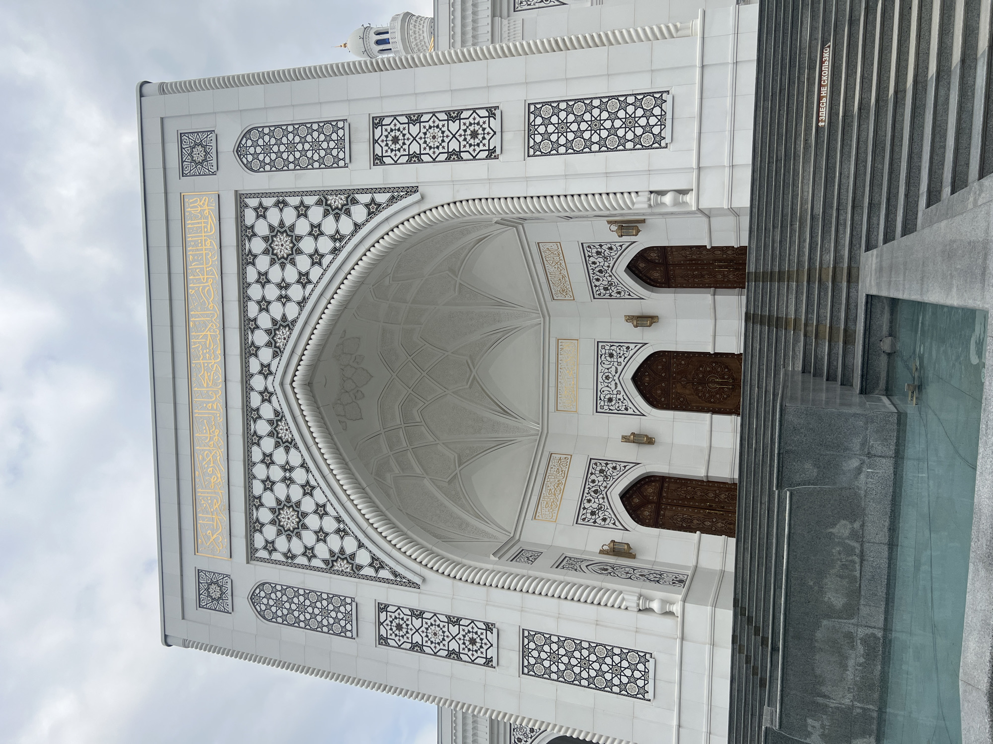 The image captures the exterior of the Akhmad Kadyrov Mosque in Shali, Russia, under a cloudy sky. The mosque is a grand, white structure adorned with intricate Islamic geometric patterns in black and gold. A prominent arched entryway features three large, ornate wooden doors. Above the entrance, golden Arabic calligraphy is displayed. The mosque is approached by a wide set of grey granite stairs, bordered by a shallow water feature. The architectural style is modern Islamic, characterized by its clean lines, decorative motifs, and prominent minaret visible to the right. There are no people visible in the image, and the scene is static and peaceful.