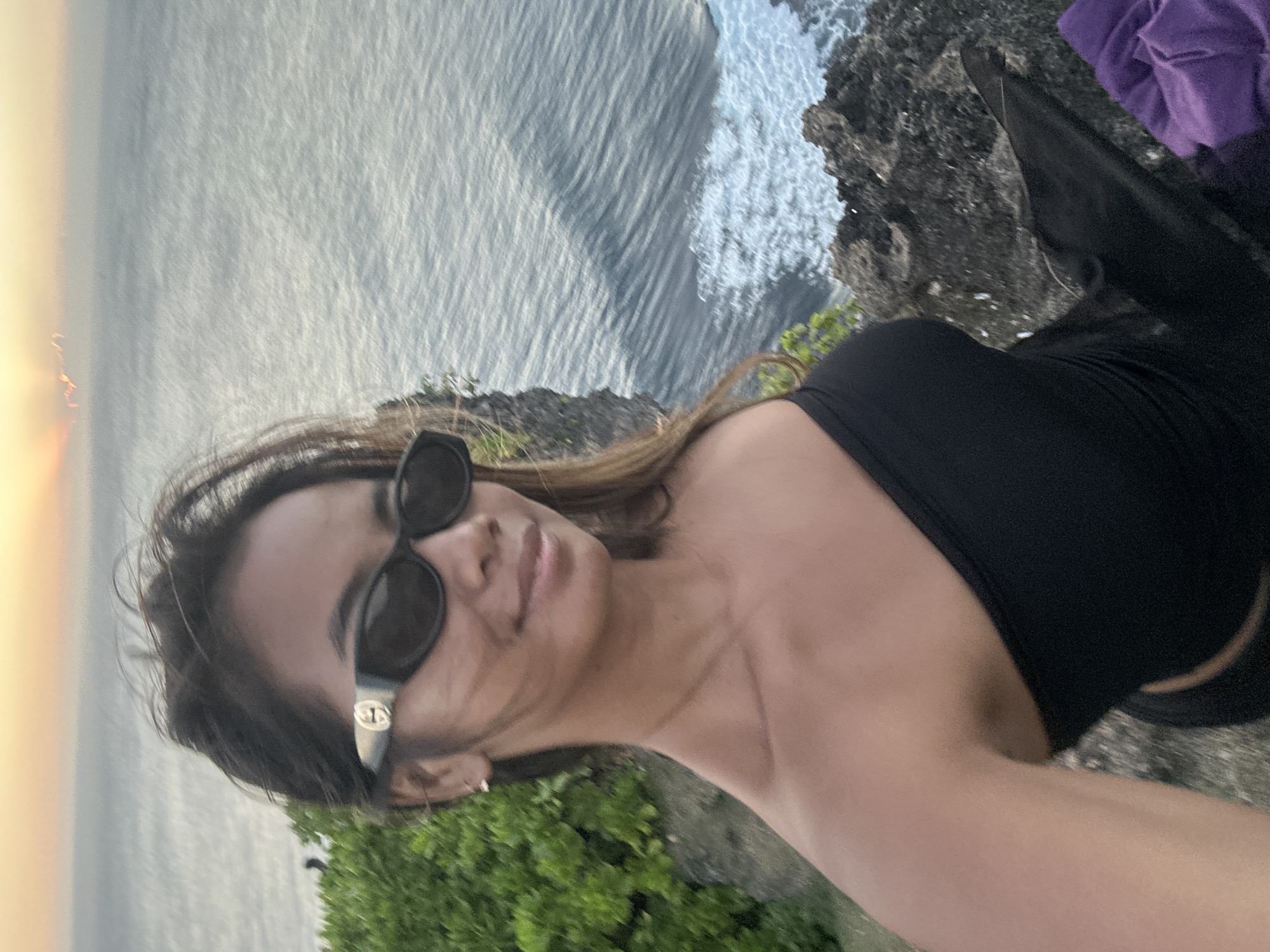 A young woman is taking a selfie on a cliff overlooking the ocean at sunset. She is wearing a black strapless top and sunglasses. The sun is low in the sky, casting a warm glow over the water. The waves are crashing against the rocks below. There is a purple cloth on the ground next to her. The location is the south coast of Bali, near Kuta Selatan, Indonesia.
