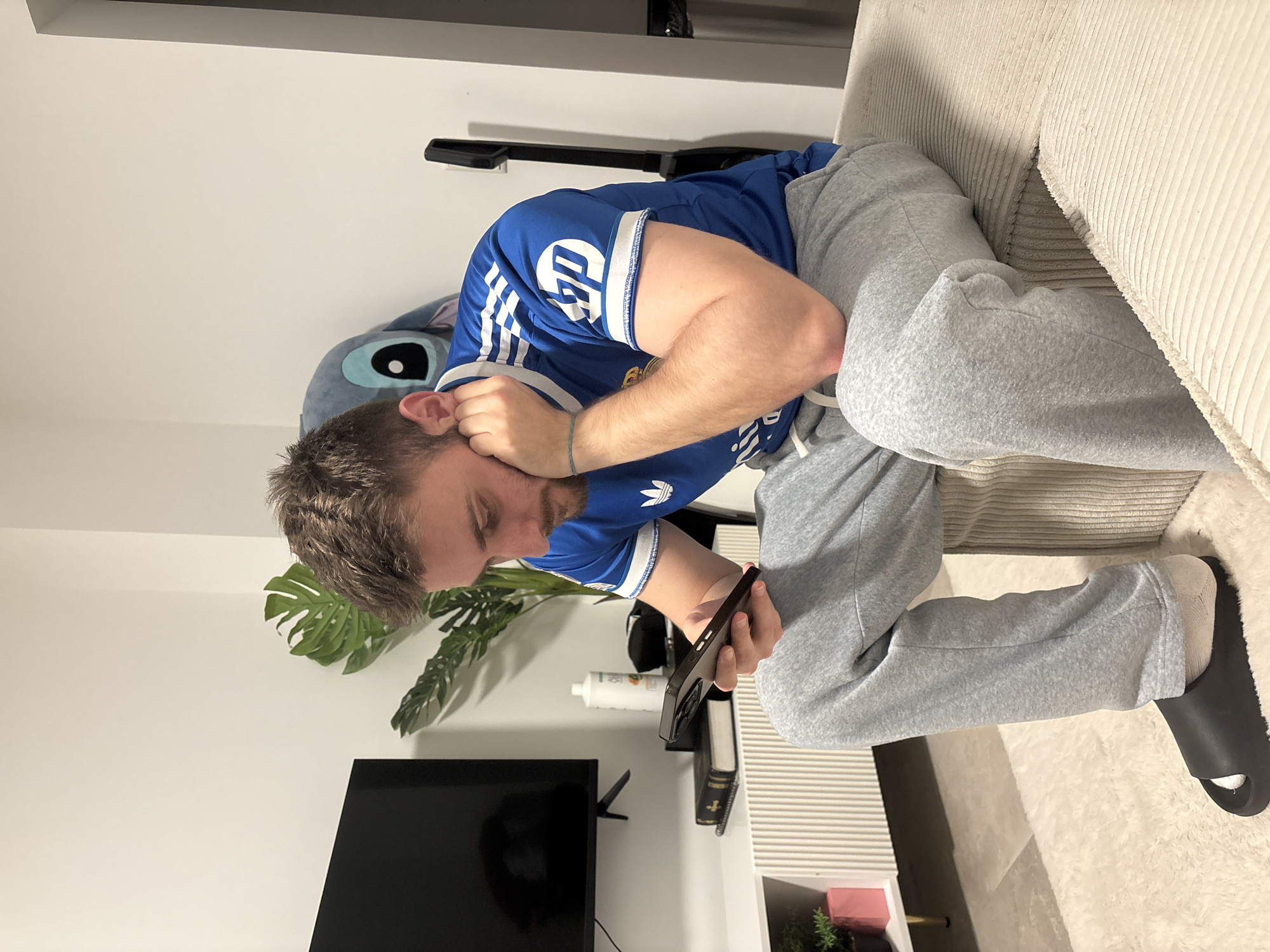 A man is seated on a beige corduroy couch, wearing a blue Chelsea FC jersey with Adidas stripes and light gray sweatpants. He is holding a black smartphone in his right hand. His left hand is near his face. He is wearing a black slide on his right foot. Behind him is a gray plush toy, a guitar leaning against the wall. A Monstera plant and a television are visible to his left, along with a white shelf unit containing a bottle of white liquid and a book. The location is possibly a living room. The information available does not allow to confirm this is in Mönchengladbach, Germany.