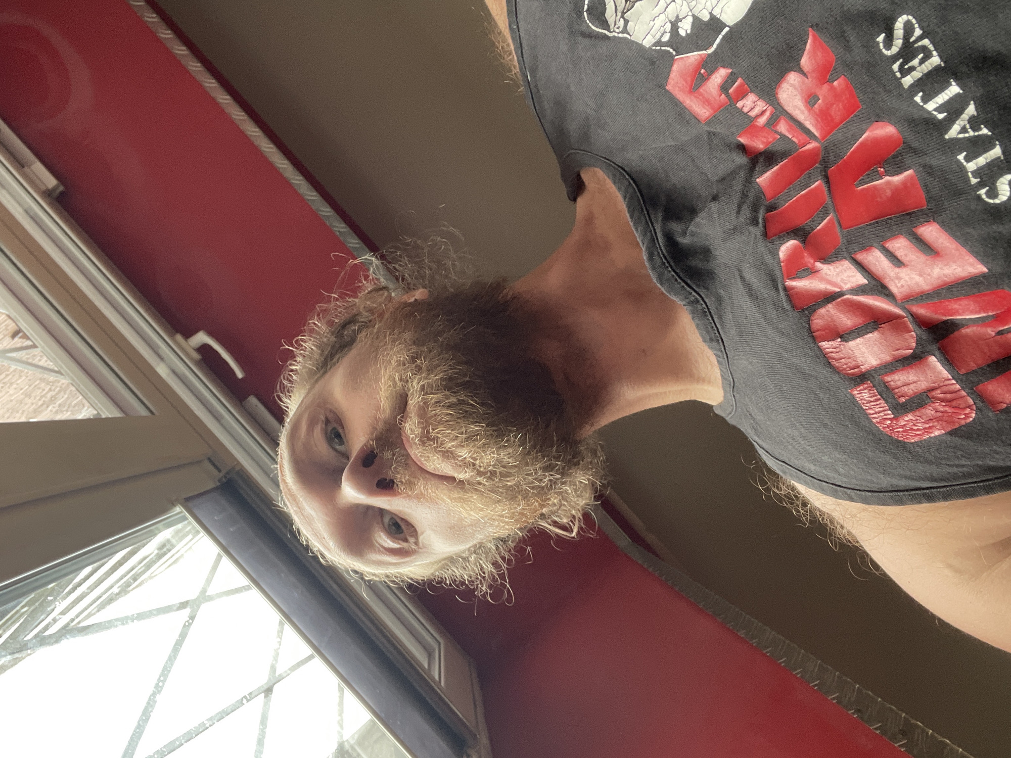 A low-angle, close-up, selfie-style shot shows a man with long, light brown, unkempt hair and a full beard. He is wearing a dark grey tank top with red lettering that says "GORILLA WEAR" in a distressed font, and also some white lettering that appears to say "STATES" below it. The man is looking directly at the camera with a neutral expression. He is positioned in front of a red surface, with a window frame visible above and to the left. The window appears to have a metal grate or structural element visible through it. The background is a muted brown. There is no discernible information in the image that places the location in Saint Petersburg, Russia.
