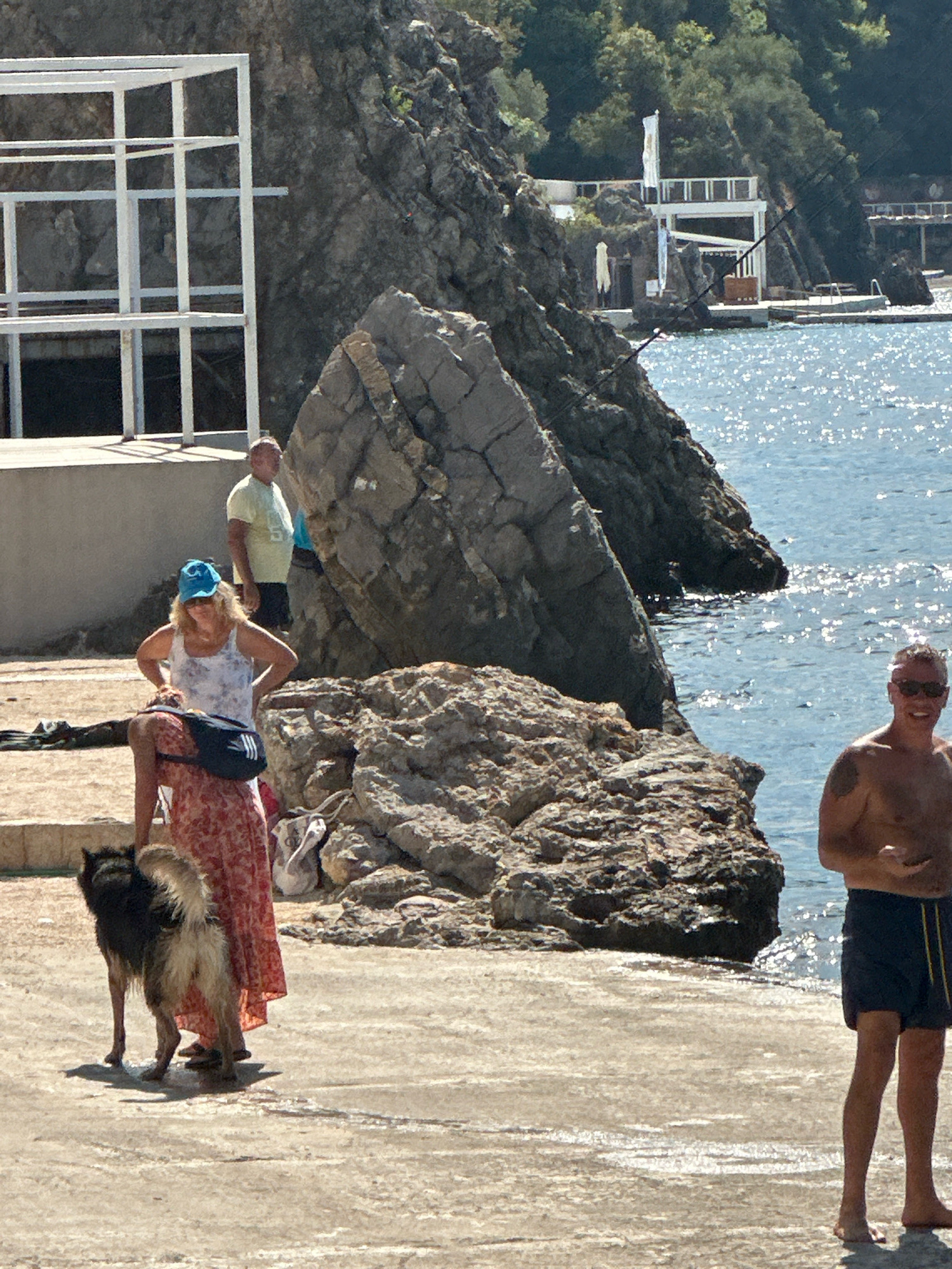 A man and two dogs are on a stone pier, with a woman standing behind them. The man, wearing sunglasses and dark blue swim trunks, is shirtless. One dog is black and tan with a long tail, and the other is partially obscured. The woman, wearing a blue baseball cap and a white and blue floral tank top with a long red and white floral skirt, has a black fanny pack around her waist. A light-colored, possibly bird-like, object rests on the pier behind them. The scene is set against a backdrop of a rocky shoreline and the Adriatic Sea, with the city of Budva, Montenegro, visible in the distance with white buildings nestled into the lush green hills. Sunlight glints off the water's surface.