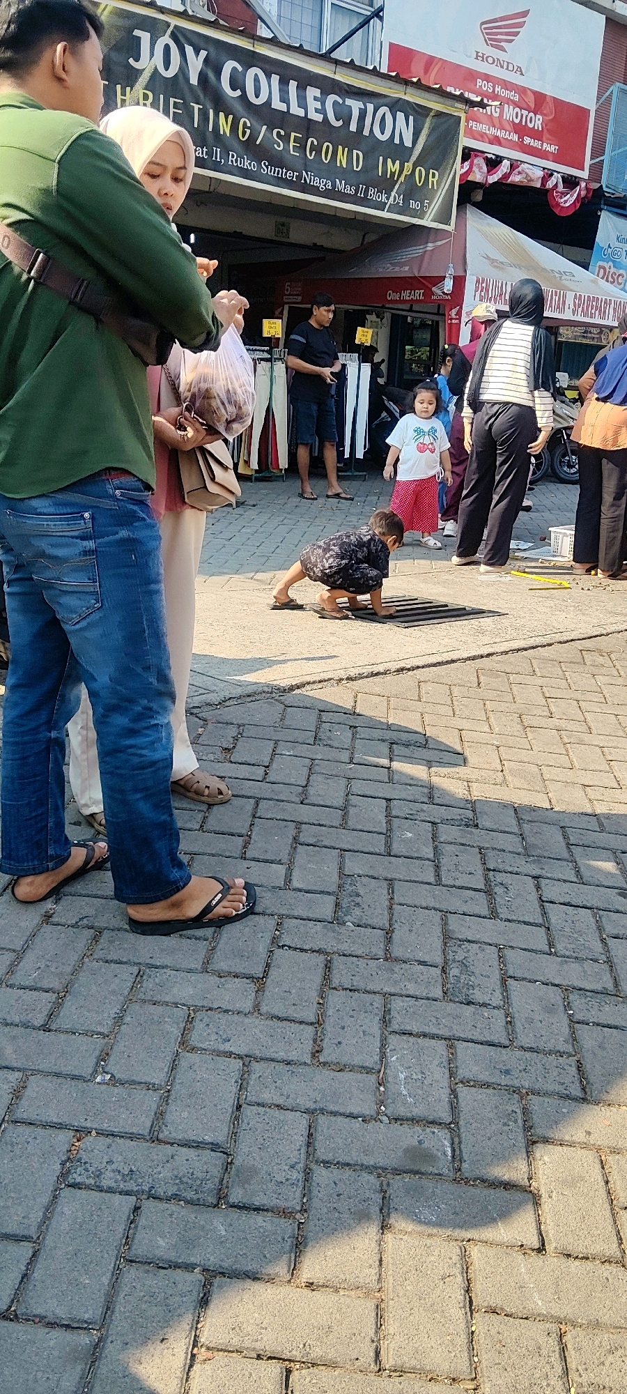 A low-angle, full-shot photograph shows a child on all fours on a patterned brick sidewalk in front of a storefront in Cibatu, Indonesia. The child, wearing a dark, patterned shirt and flip-flops, is reaching towards a dark grate on the ground. The child's legs and feet are visible, as are the bottom of a woman's legs in a pink headscarf and beige pants, and a man's legs in blue jeans and flip-flops, standing to the left. In the background, there are people standing and walking, as well as several storefronts. A red and white sign above one of the storefronts reads "HONDA." Another sign reads "JOY COLLECTION" and "THRIFTING/SECOND IMPOR." The lighting suggests it is daytime, with shadows cast across the sidewalk.