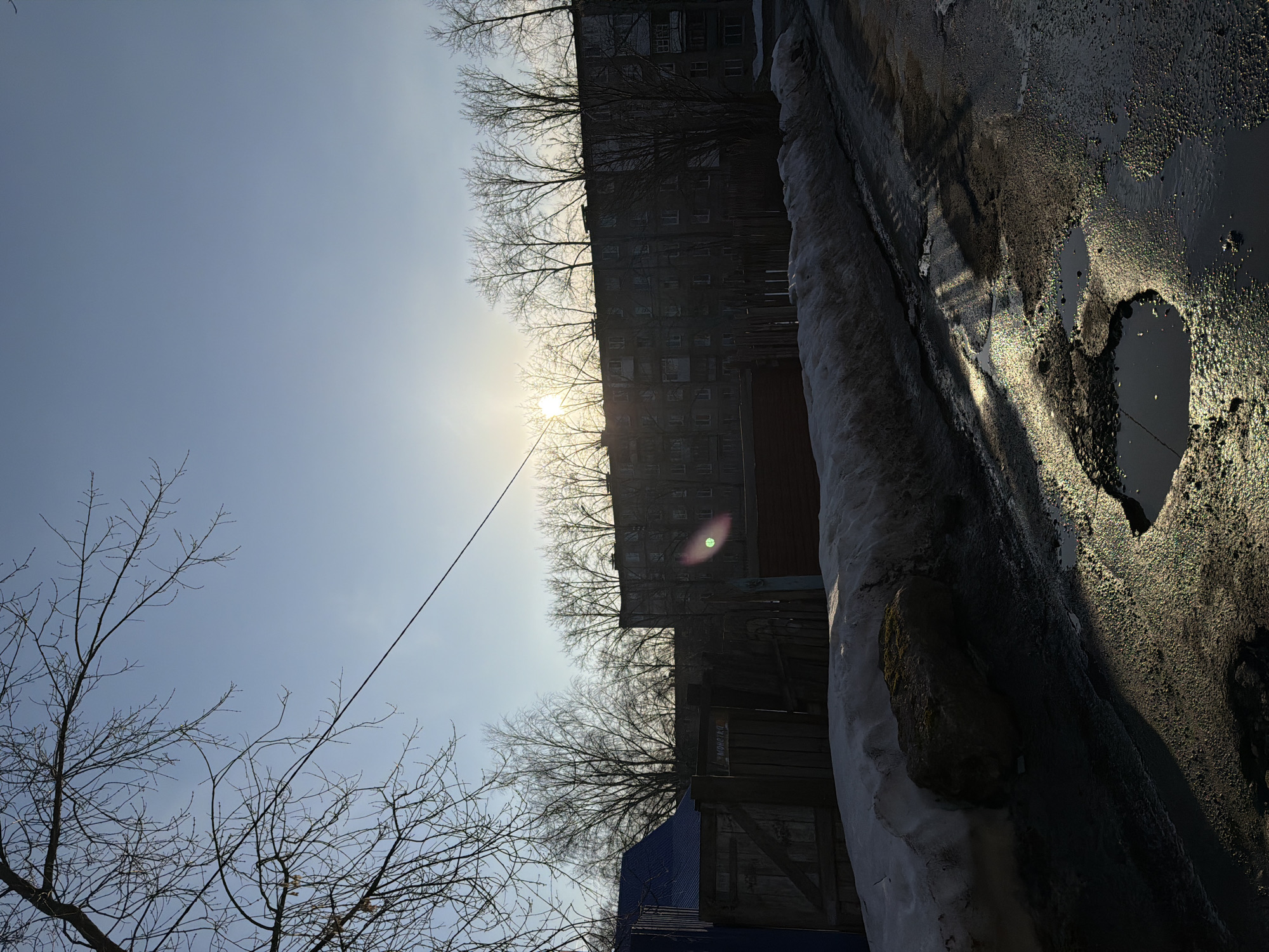The image, taken in Berezniki, Russia, presents a view of a winter landscape. A building with numerous windows occupies the central horizon line, silhouetted against a bright sun. Bare trees line the top and bottom of the building. A utility wire stretches across the sky. In the foreground, there is a snowy and icy surface with a puddle, reflecting the light. There is also what looks like a rock or debris on the side of the snow. Portions of the infrastructure include parts of a building in need of repair.