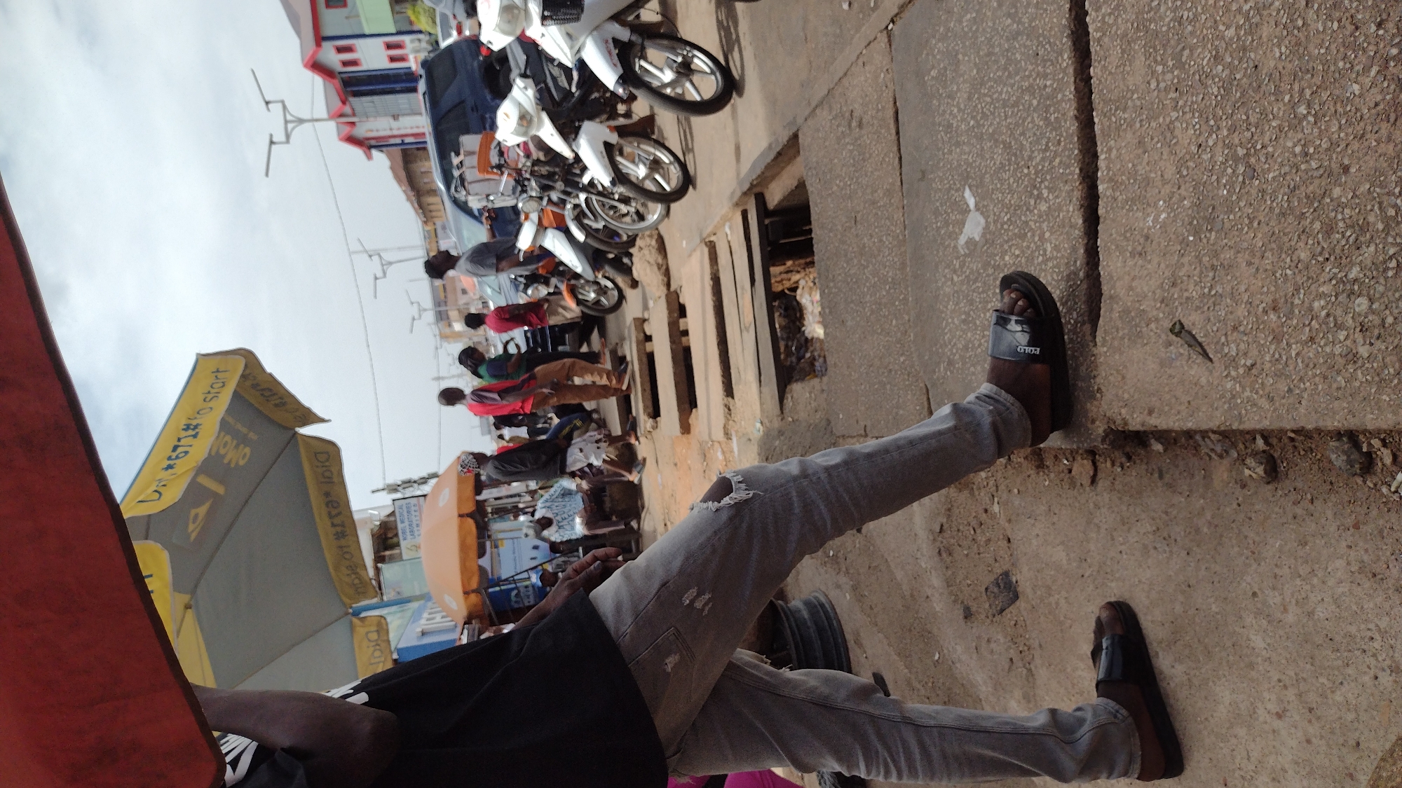A street scene in Jos, Nigeria, is viewed from a low angle. A person wearing grey jeans and black sandals is positioned in the foreground, extending across the frame. The person also appears to be wearing a black shirt. In the background are several motorcycles parked along the side of the road, with people gathered nearby. Adjacent to the motorcycles is a storm drain. A row of market umbrellas and canopies line the street. The buildings in the distance are a mix of low-rise commercial structures. The sky is overcast.