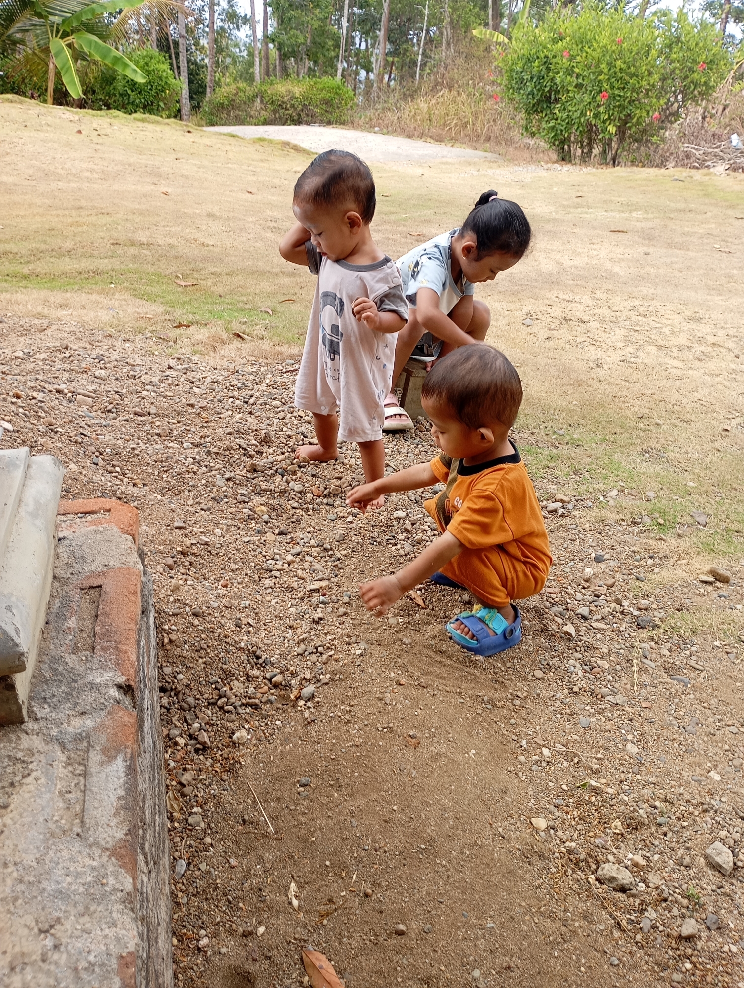 Three young children are gathered on a patch of dry, sandy ground with scattered pebbles and sparse grass, near a low, weathered stone structure. The child in the foreground, wearing an orange outfit and blue sandals, is crouched down and reaching towards the ground. To the left, another child in a light-colored dress stands, looking down and holding a hand to their ear or head. In the background, a third child, with dark hair pulled into a ponytail and wearing a light blue and white shirt, is also crouched, appearing to be looking at something on the ground. Behind them, a dry, grassy slope rises towards a line of trees and shrubs. The scene suggests an outdoor, possibly rural or semi-rural, setting in Ketepung, Indonesia, with the children engaged in exploring or playing.