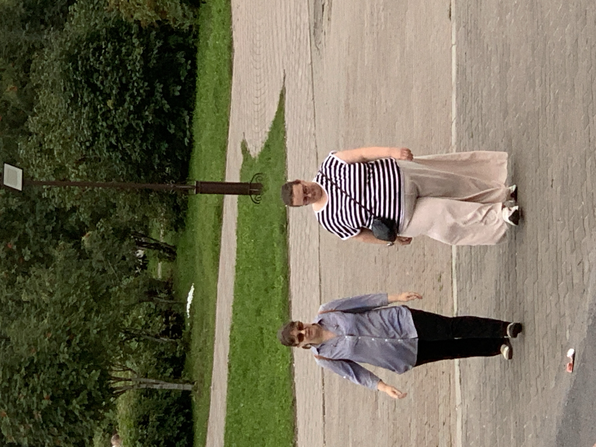 Two women walk on a paved walkway in Prokopyevsk, Russia. The woman on the left wears a blue collared shirt and black pants, with sunglasses and a brown backpack. The woman on the right wears a black and white striped shirt and light-colored wide-leg pants, with a black crossbody bag. Behind them, a street lamp stands near a grassy area and trees. The ground is paved with cobblestones, and some litter is visible on the walkway.