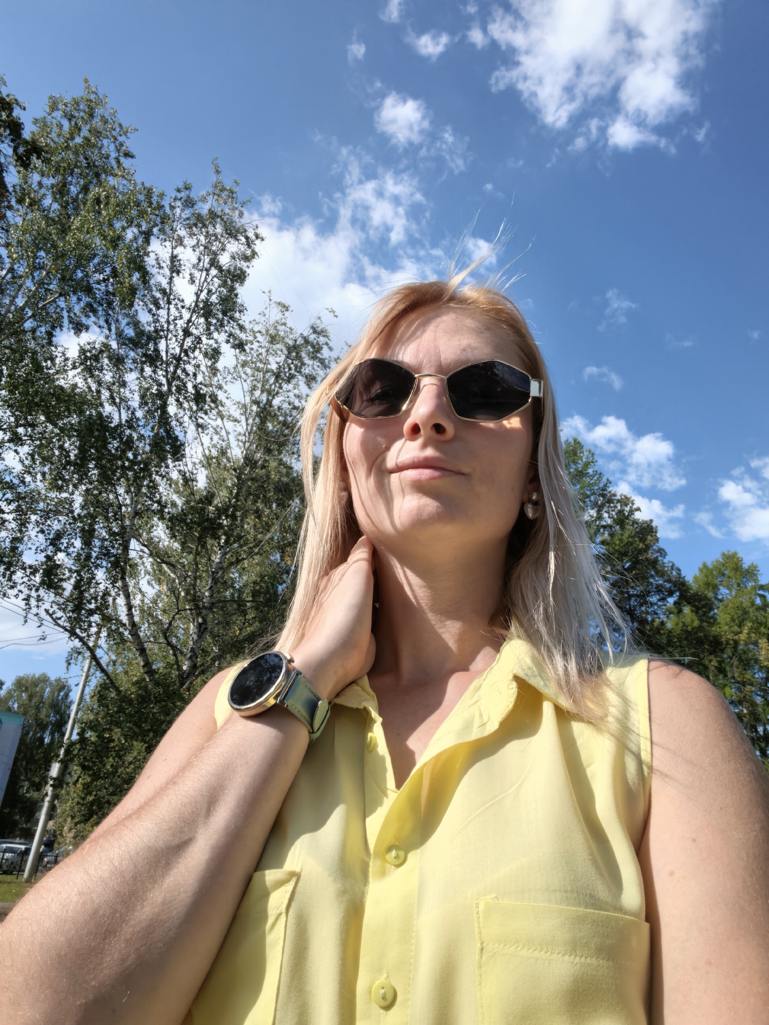 The image is a selfie of a Caucasian woman with blonde hair and sunglasses, taken outdoors with a bright blue sky and fluffy white clouds in the background. She is wearing a yellow sleeveless shirt with buttons. The woman's left arm is raised, and she is touching her neck with her left hand. She is wearing a round smartwatch with a green band on her left wrist. Trees with green foliage are visible behind her, as well as a glimpse of a building and a parked car on the left side of the image. The scene appears to be a sunny day in a park or residential area. Based on the provided information, the location is identified as Sterlitamak, Russia.