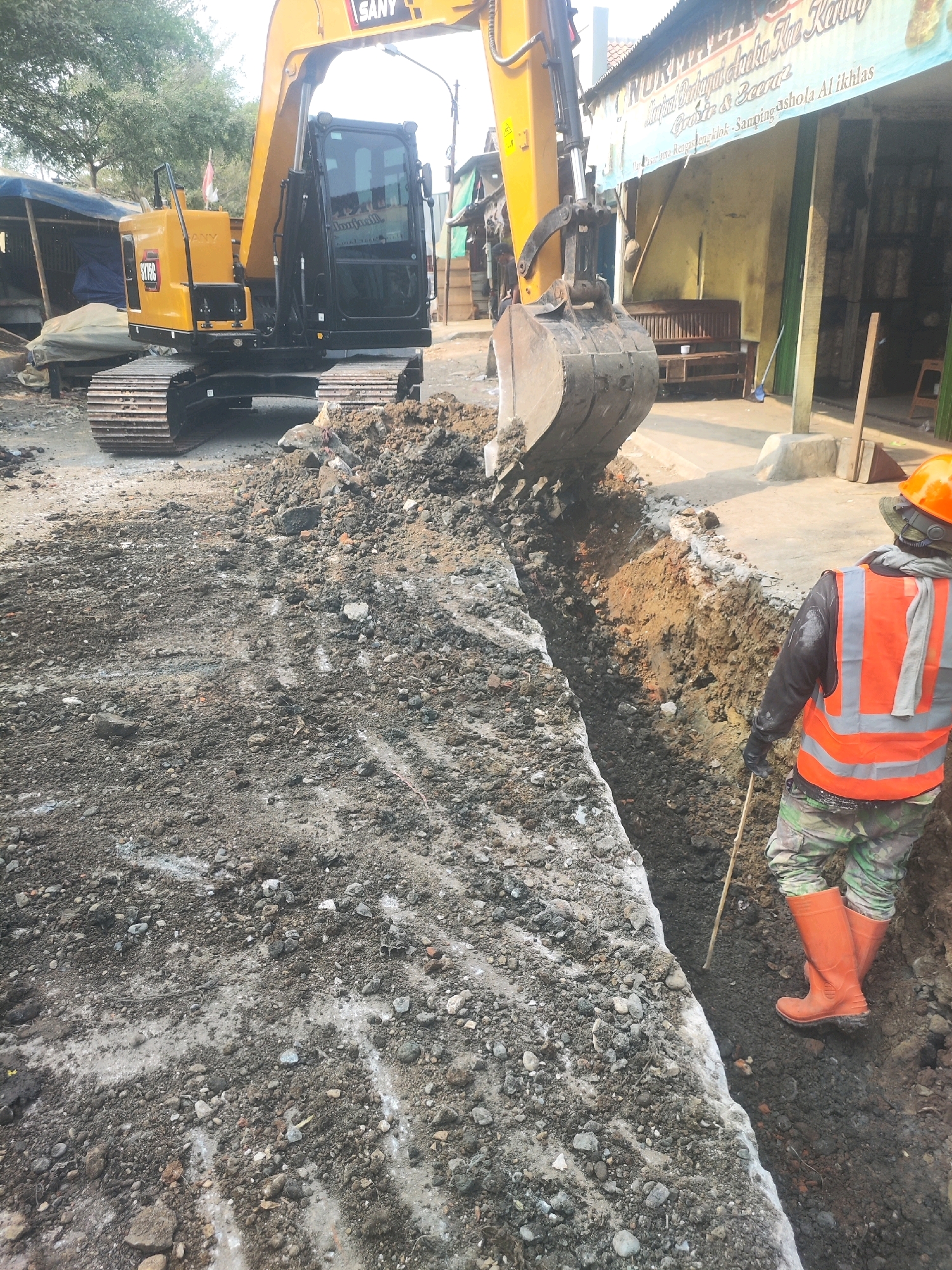 The image depicts an outdoor construction scene, likely in Karawang, Indonesia. A large yellow excavator is parked on the left, its bucket partially filled with dirt. In the foreground, a trench has been dug in the ground, revealing layers of soil. A worker, wearing an orange safety vest, camouflage pants, and orange boots, is standing on the right side of the trench, holding a long stick, possibly inspecting the trench or directing operations. Behind the excavator, a building with signage is visible, suggesting a commercial or residential area. The ground is uneven and covered with dirt, rocks, and debris, characteristic of a construction site. The overall atmosphere suggests ongoing infrastructure work.