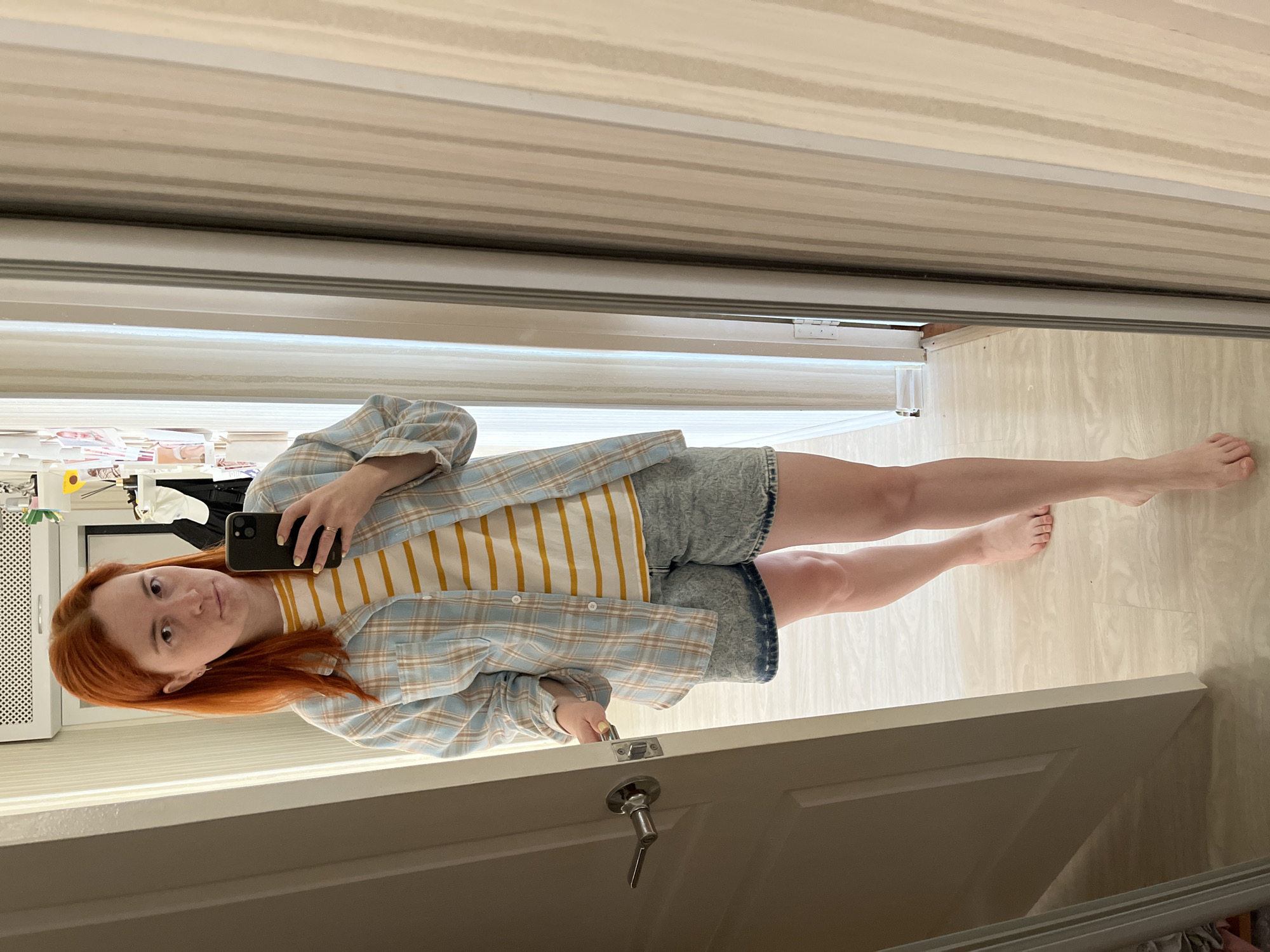 A Caucasian woman with shoulder-length, bright orange hair is standing in what appears to be a bedroom, possibly in Chaikovsky, Russia, based on the provided information. She is taking a mirror selfie, holding a black smartphone in her right hand. She is wearing a yellow and white striped t-shirt layered under an open blue and white plaid shirt, and distressed denim shorts. Her left hand is near the door handle of an open door. The floor is light wood laminate. In the background, a wall with vertical striped wallpaper is visible, along with a shelf displaying various items, including photographs.