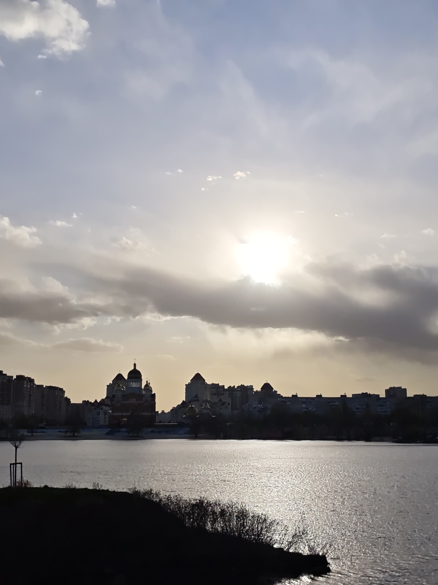 The image presents a serene cityscape viewed across a body of water, likely a lake or river, at what appears to be late afternoon or early evening. The bright sun is positioned centrally in the upper sky, partially obscured by a horizontal band of darker clouds, while lighter, scattered clouds populate the rest of the blue-grey sky, which takes on a warm, golden hue near the horizon.
Across the shimmering water, the city skyline is silhouetted against the bright sky. A prominent Eastern Orthodox church or cathedral, characterized by its multiple domes, some topped with crosses, stands out as a central architectural feature within the silhouette. This architectural style is consistent with the provided location context of Kyiv, Ukraine. Various other buildings form the dense cityscape beyond the water.
In the immediate foreground, a dark, silhouetted landmass with sparse bushes creates a shoreline. On the left side of the foreground, a bare, abstract, tree-like structure or sculpture is visible. The scene is devoid of people or direct activity, focusing instead on the dramatic lighting and the urban landscape. There is no visible text in the image.