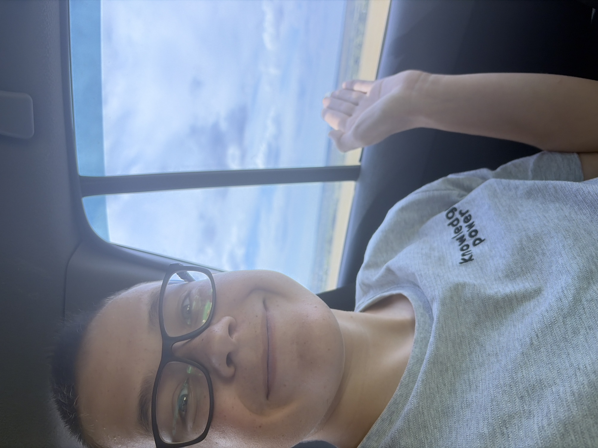 A close-up shot captures a person inside a vehicle, their face turned towards the camera. The person is wearing black-rimmed glasses and a grey t-shirt with the embroidered text "knowledge power" visible on the left chest. Their right hand is raised, fingers slightly spread, as if waving or gesturing. Outside the car window, a landscape unfolds under a cloudy sky. The image appears to be taken in Novoe Teneevo, Russia.