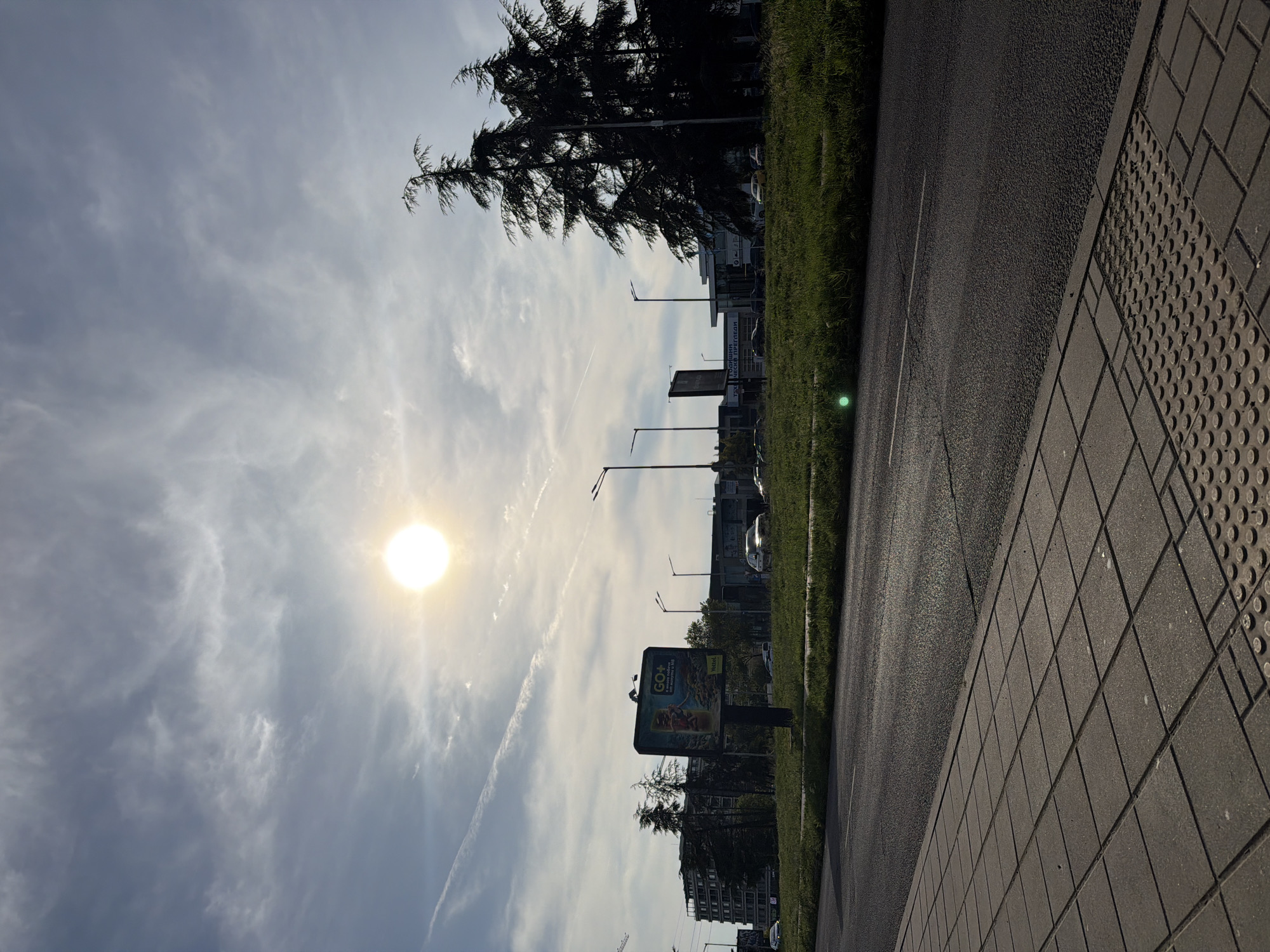 The image depicts an outdoor scene with a view oriented towards the sky and a street. The sun is visible through the clouds in the upper left part of the image. A contrail runs diagonally across the sky.
The right side of the image shows a paved walkway, followed by a road and then a grassy area. Beyond the grass is a line of trees and buildings. Lampposts are visible along the road. A large advertisement board can be seen amongst the other objects.
Given the advertisements, the buildings, and the vegetation, the image is likely taken in a city area. I lack specific information to confirm but based on the hints provided, it's in Burgas, Bulgaria. There are no people visible in the image.