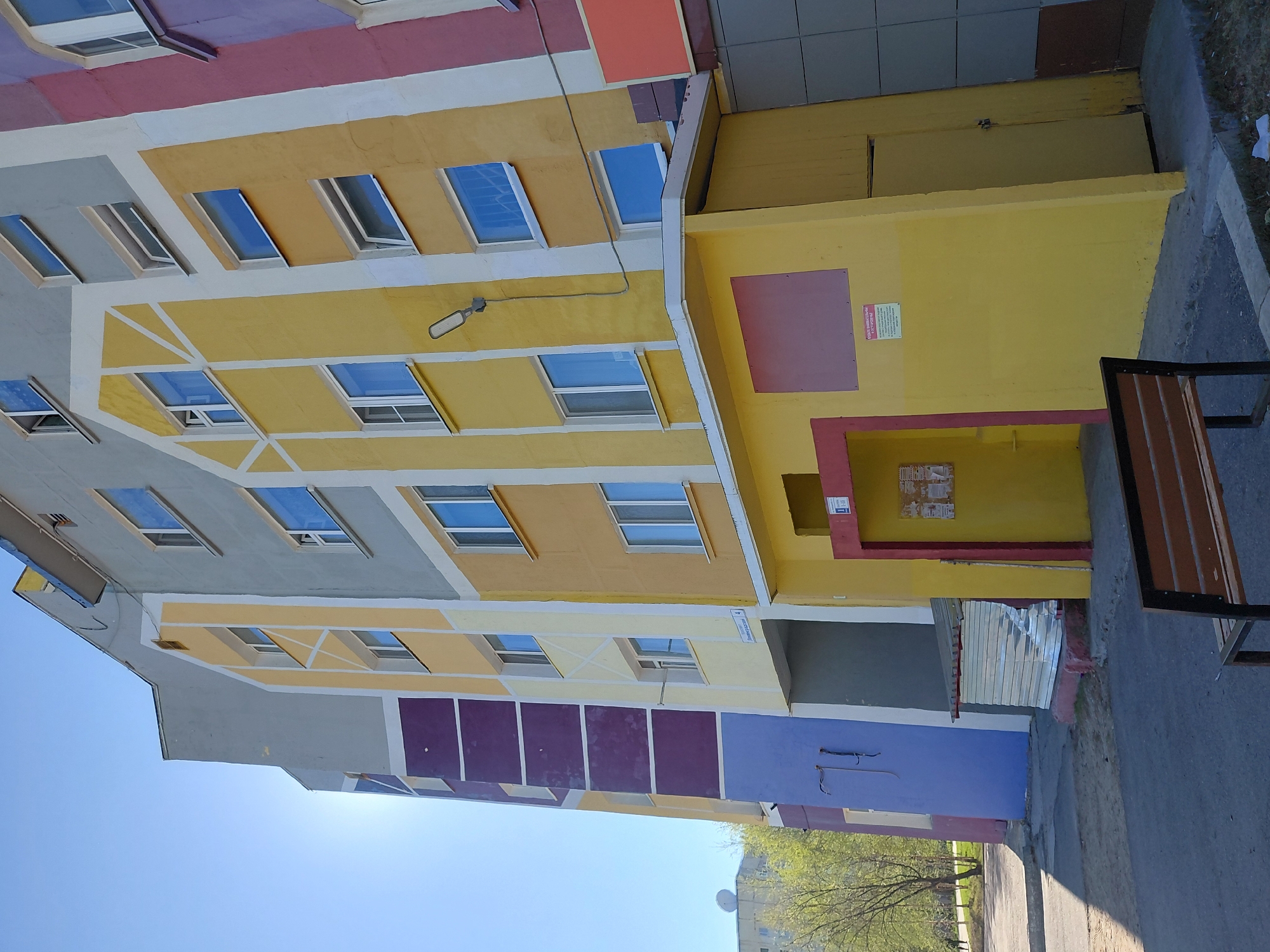 The image captures a vibrantly painted apartment building under a clear blue sky on a sunny day in Nadym, Russia. The multi-story building features a facade divided into distinct sections of bright yellow, light gray, purple, and a reddish-pink color along the upper edge and roofline. Numerous rectangular windows, many resembling dormer windows, are present across the different colored sections, with some appearing to be slightly ajar. A white drainpipe runs down the building, and an elongated white streetlight is mounted on a yellow section of the wall.
At ground level, a smaller, single-story yellow structure is attached to the main building, featuring a recessed entrance framed in red. Above this entrance, a reddish-pink rectangular sign displays the Russian word "ОСТОРОЖНО" (CAUTION), followed by smaller, unreadable warning text. To the right, a wooden and metal bench sits on a concrete sidewalk. Further along the building's base, a silver rolling shutter is visible near a purple-painted section, suggesting a commercial or service entrance. A small white sign on the yellow wall clearly indicates the address: "Проспект Ленина, 4" (Lenin Avenue, 4). Other posters are affixed to the wall near the shutter, but their text is illegible.
Green trees and a small white satellite dish are visible in the distance on the left side of the frame. The strong sunlight creates clear shadows, emphasizing the bright colors of the building. There are no people visible, and no specific activity is taking place; the scene depicts a static view of the building and its immediate surroundings.