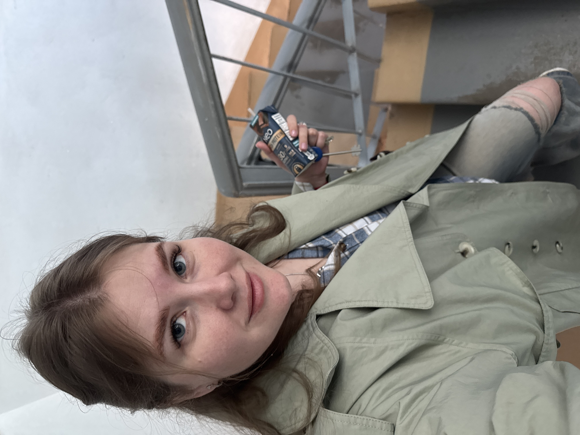 The image is a selfie taken by a young woman with light brown hair and blue eyes. She is wearing a light green trench coat over a blue and white plaid shirt and ripped jeans. She is holding a small carton of "neo protein" drink in her right hand. The background shows a staircase with metal railings and steps alternating between gray and beige colors. The location is likely in the city of Perm, Russia, inferred from the drink packaging (presuming availability reflects the geographical location).