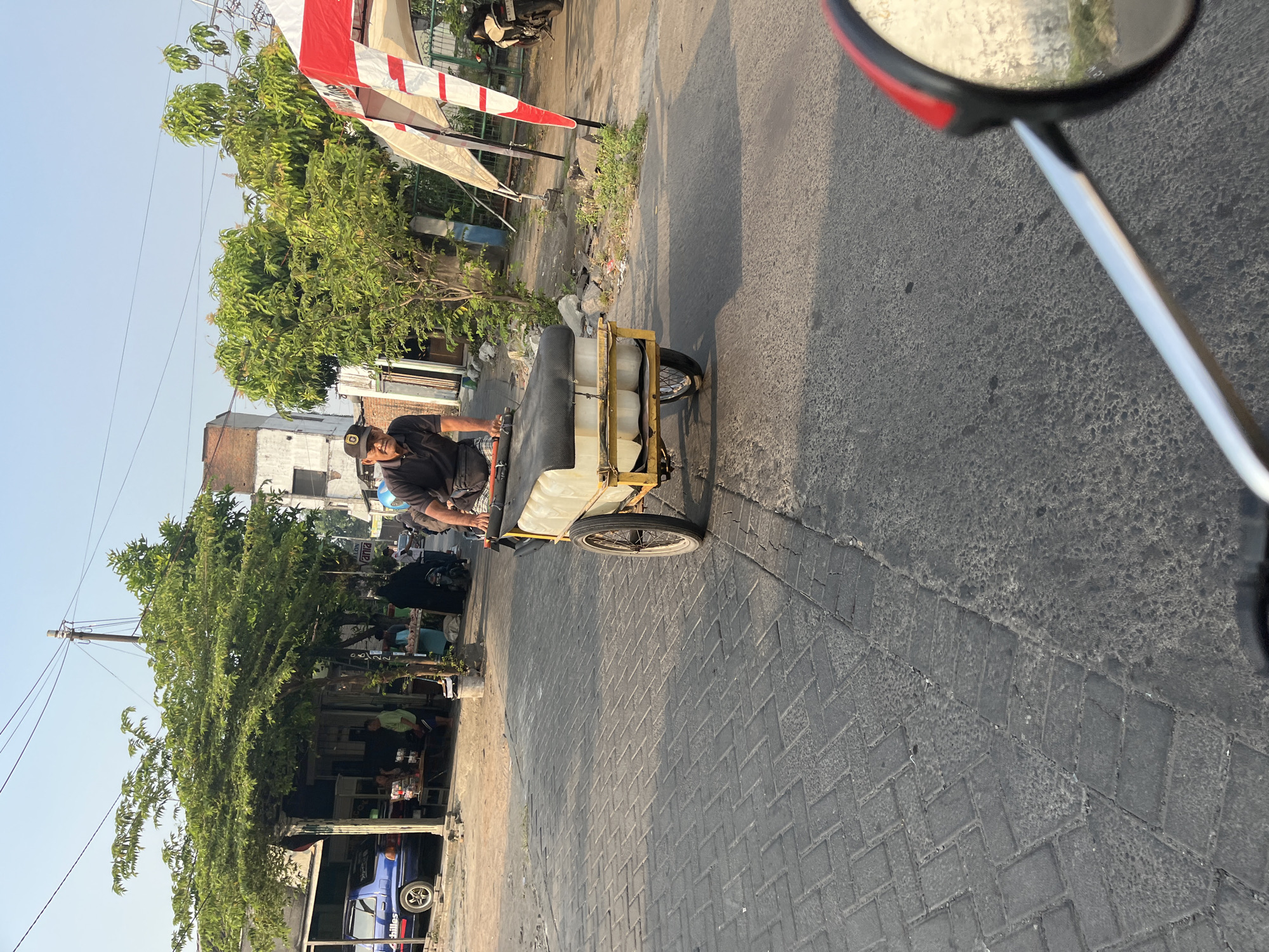 A man wearing a black polo shirt and a black baseball cap rides a three-wheeled cargo bicycle in Tenggulunan, Indonesia. The bicycle is loaded with what appear to be two large white containers. The man is looking forward and appears to be pedaling. The bicycle is on a paved road. In the background, there are buildings, trees, and other vehicles, including a blue car and a motorcycle. There are also people standing near a food stall on the side of the road. The scene is illuminated by the warm light of what appears to be late afternoon or early morning.