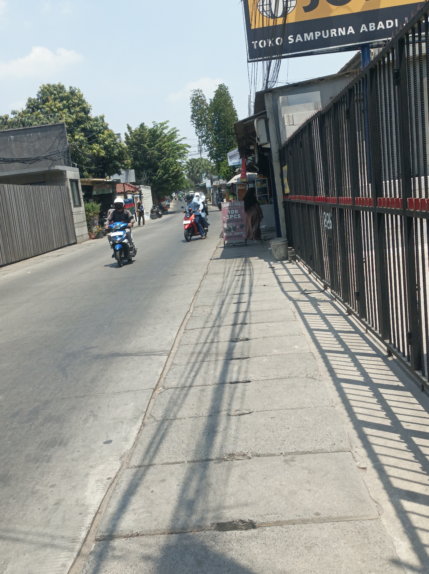 The image depicts a street scene in West Jakarta, Indonesia. A road runs diagonally from the lower left to the upper right of the frame, with a concrete sidewalk on the right side. Several individuals are riding motorcycles on the road. In the foreground, a person on a blue motorcycle is approaching the viewer. Further down the road, another person is on a red motorcycle. On the right side of the sidewalk, a black metal fence with red accents runs along the edge of a property. Behind the fence, a building with a sign that reads "TOKO SAMPURNA ABADI" is visible. The sky is clear with some scattered clouds. The lighting suggests it is daytime.