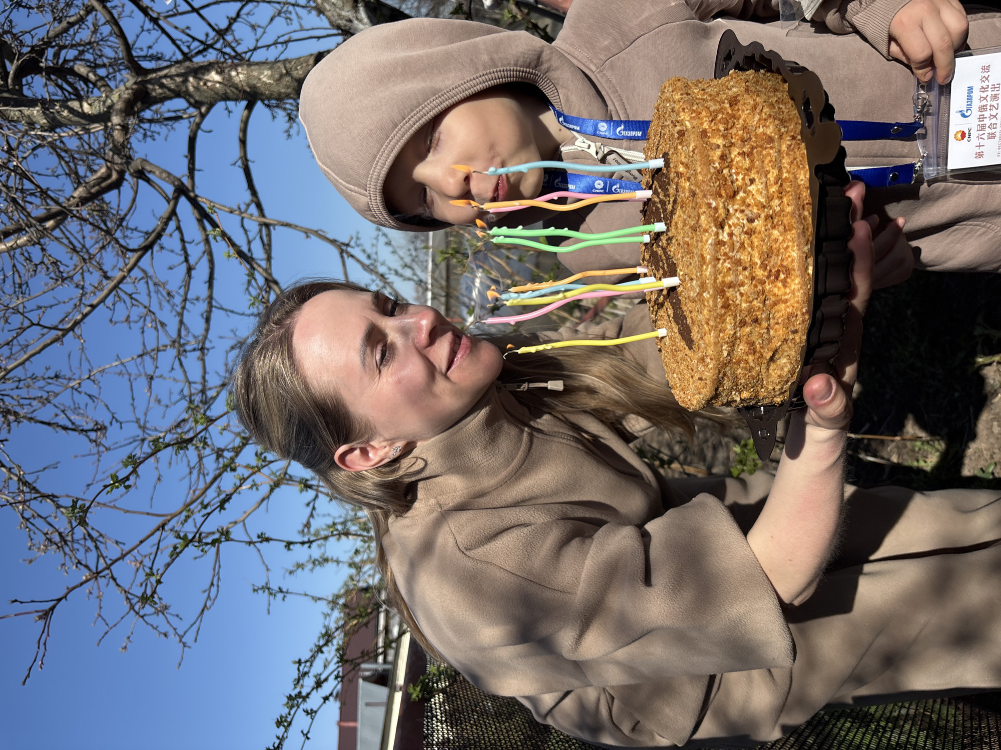 A medium shot captures a woman and a child holding a cake with lit candles outdoors. The woman, fair-skinned with blonde hair, wears a tan jacket and looks at the child. The child, hooded in a tan sweatshirt, leans towards the cake, possibly about to blow out the candles. The cake is round, topped with a crumbly layer, and has several brightly colored candles inserted into it. Both individuals are wearing lanyards with the same badge attached.
The background reveals a tree with bare branches and a clear blue sky. There are elements suggesting an urban setting, possibly a park or residential area in the city of Chaikovsky, Russia, given that the badges appear to be in Russian. A fence is visible in the lower left of the frame. The primary action is the holding of the cake and the anticipation of blowing out the candles, which implies a birthday or celebration.