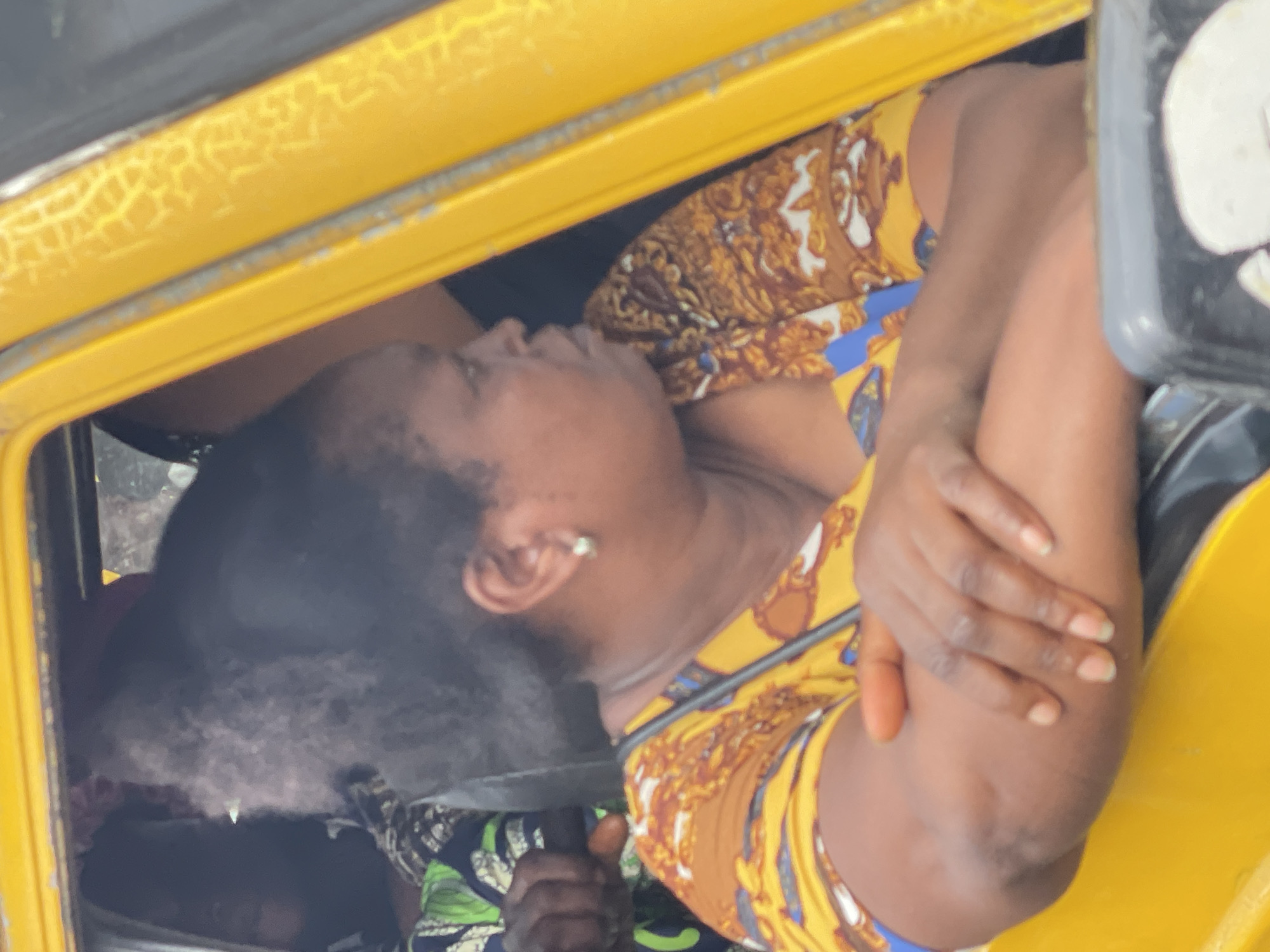 An unfiltered, medium shot shows a Black woman in profile, looking to her right, inside the yellow interior of a vehicle. She has dark, curly hair pulled back into a bun, and she wears a pearl earring. Her arms are crossed in front of her, and she is dressed in a colorful yellow and brown patterned top. The interior of the vehicle appears to be a bus or taxi, with a yellow frame visible on the right side of the image. Another person is visible in the background to the left of the woman, wearing a green and black patterned garment. The woman's gaze suggests she is observing something outside the vehicle, possibly the busy streets of Onitsha, Nigeria, a bustling commercial hub known for its vibrant markets and traffic congestion.
