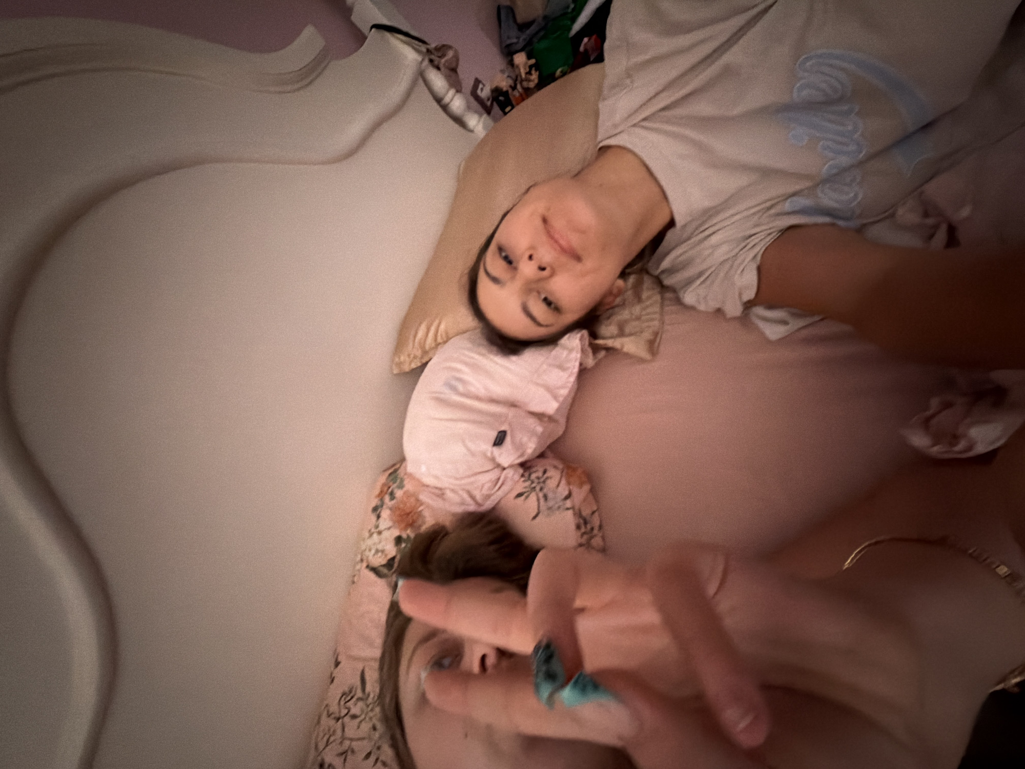 Two young women are lying in a bed, with the camera in the foreground. One woman is closer to the camera, with her face turned towards it and her right hand raised, making a peace sign. She has painted blue fingernails. The other woman is lying further back, with her head on a pillow. She is wearing a light-colored t-shirt with blue lettering. The bed has pink sheets and a floral patterned pillow. The environment appears to be a bedroom, with a headboard visible in the background. The image was taken in Tsarevo, Bulgaria.
