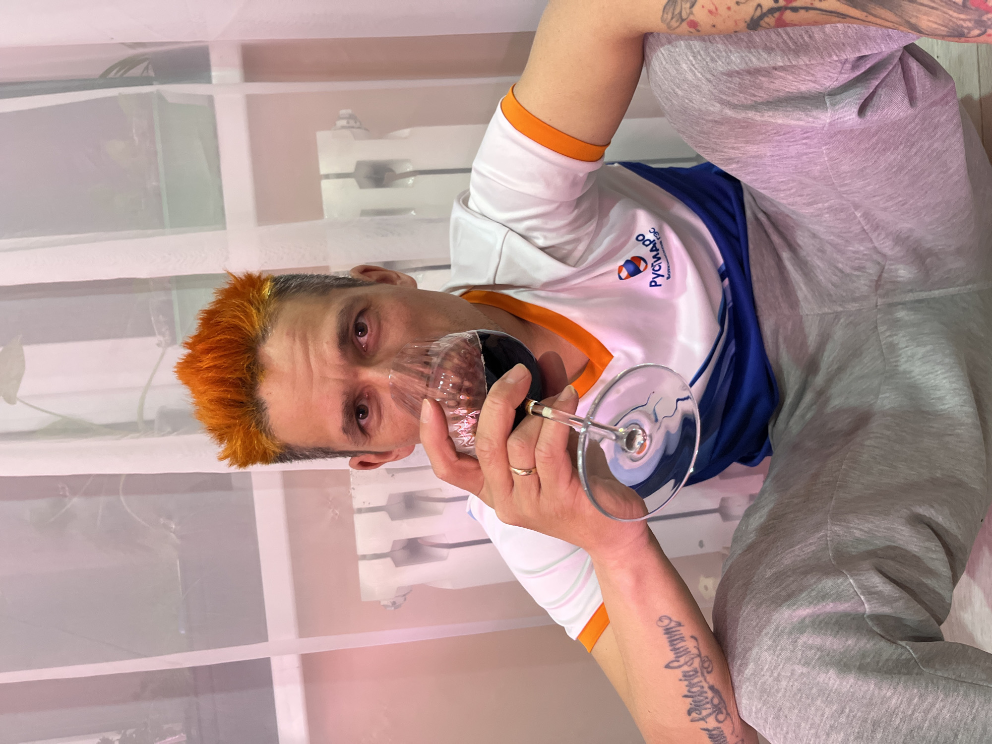 A man with orange hair is holding a glass of red wine. He is wearing a white t-shirt with blue and orange trim and gray sweatpants. He has tattoos on his arm. The background appears to be a white wall with shelves. The shirt has the name "РУСЧИМАРПО" on it, indicating the location of Chaikovsky, Russia.