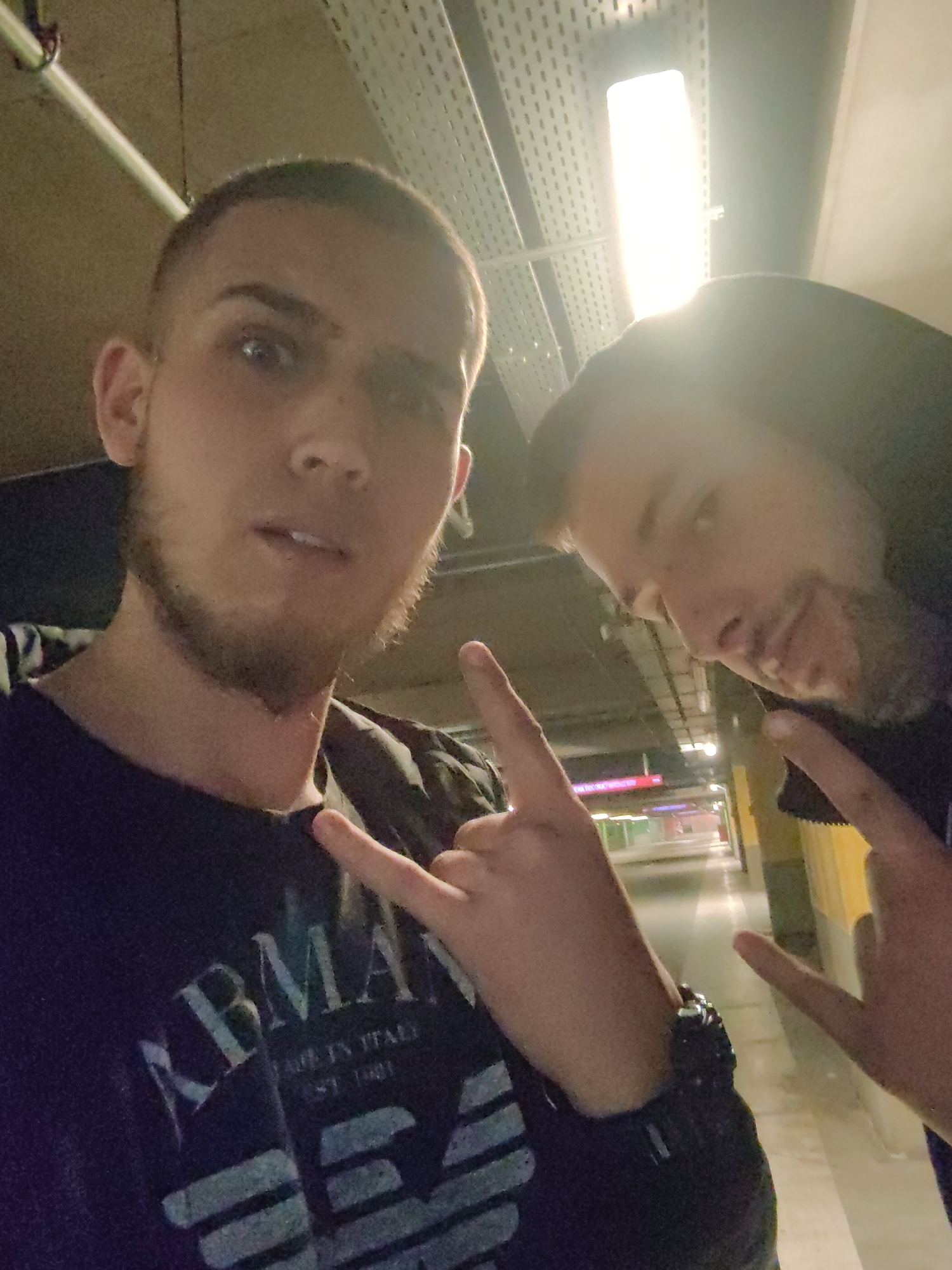 Two men pose in what appears to be an underground passage. The man in the foreground has short hair, stubble, and wears a dark t-shirt with the word "ARMANI" printed on it. He is making a "rock on" hand gesture. The second man stands slightly behind and to the right, wearing a hooded jacket or sweatshirt with the hood up. He is also making the same hand gesture. The background shows a long corridor with lights and a somewhat blurred view of possible shops or advertisements. The scene's location is not evident from the image to be specifically identified as Sofia, Bulgaria.