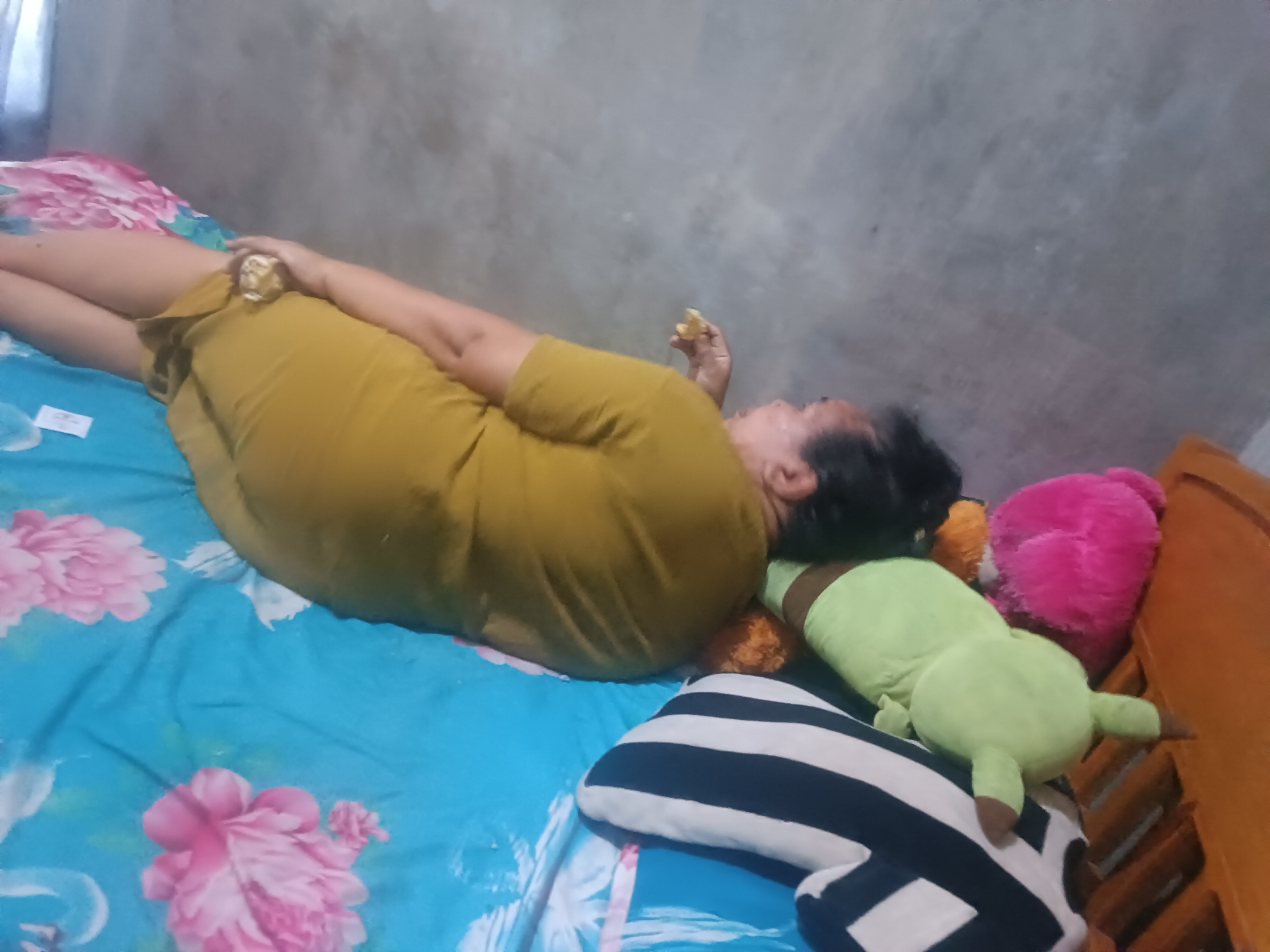 The image shows a person lying on a bed with a blue floral-patterned sheet. The person is wearing a mustard-yellow dress and is holding a piece of food in their right hand, near their mouth. There are several stuffed animals on the bed: a green one, a pink one, and a striped black and white one. The background is a plain gray wall. The scene is indoors, possibly in a bedroom. The location is identified as Tabanan, Indonesia.