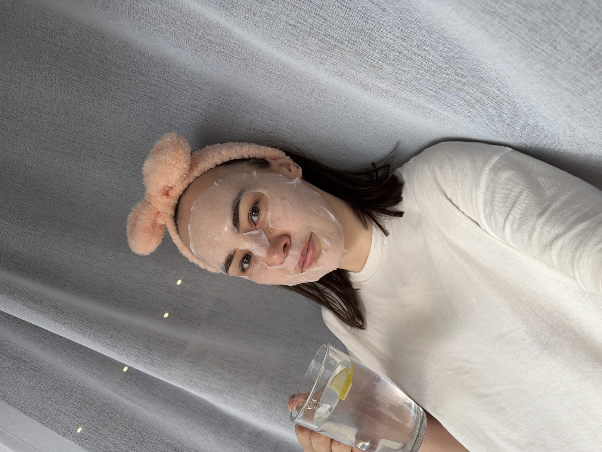 A woman is wearing a sheet mask and a peach-colored headband with bunny ears while holding a glass of water with a lemon slice in it. She is wearing a white t-shirt. The background is a grey curtain with small lights woven through it. The location is not identifiable as Chaikovsky, Russia based on the image.