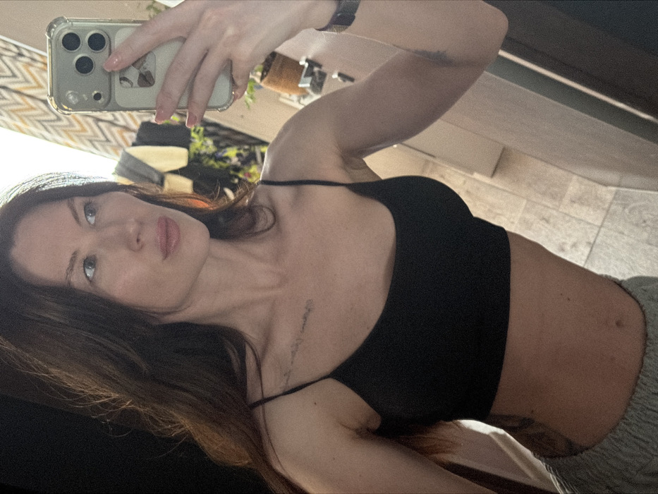 This image depicts a mirror selfie of a woman taken indoors in Perm, Russia. The subject has long, dark hair and fair skin, with her head turned slightly to the right, gazing off-frame. She is dressed in a black, thin-strapped crop top and light grey athletic bottoms, exposing her midriff. A faint linear tattoo is present on her upper left chest. A darker, larger tattoo is partially visible on her lower right torso. Her right hand, displaying light pink nail polish, holds a light-colored smartphone encased in clear material, featuring a triple rear camera array and a small white square attachment on its lower back. The background, visible in the mirror's reflection, includes a patterned curtain with a zigzag design, several hanging garments, a potted plant in a brown container, and light-colored tiled flooring.