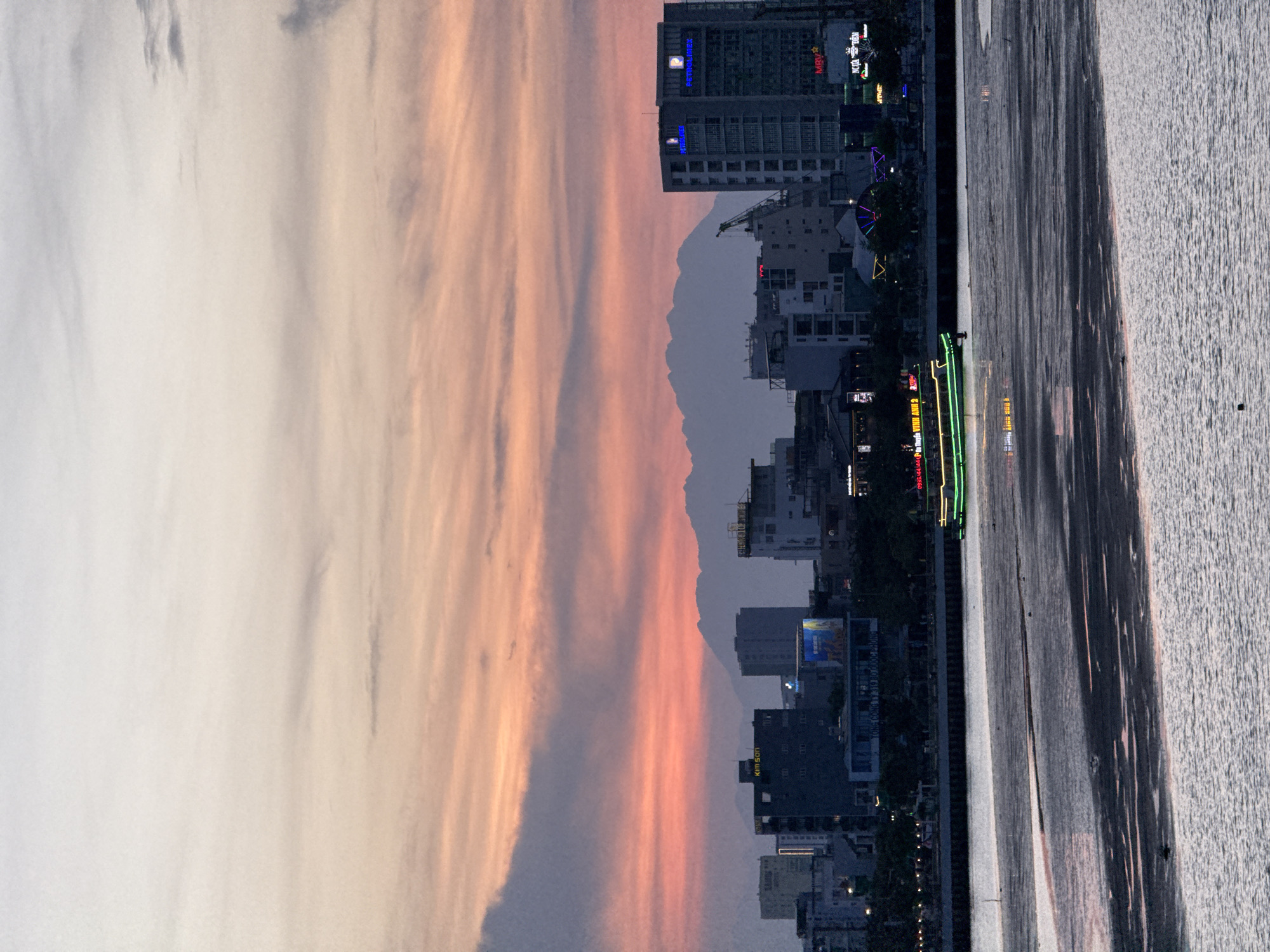 A wide shot captures a sunset scene over the Han River in Da Nang, Vietnam. The sky is painted with streaks of orange, pink, and purple, with a mountainous silhouette in the background. A modern cityscape lines the riverbank, featuring various buildings, including high-rises with illuminated signs. A green-lit boat navigates the river, while the foreground shows the textured, shallow riverbed exposed by low tide. The overall atmosphere is tranquil and visually striking, highlighting the blend of natural beauty and urban development in Da Nang.