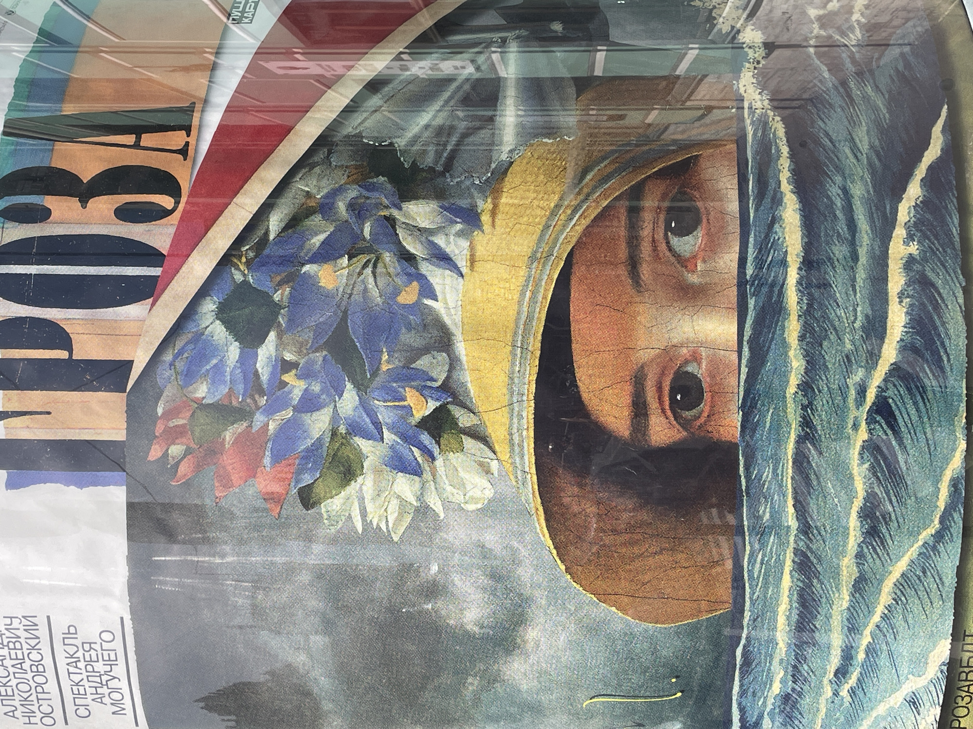 The image is a close-up of overlapping posters or advertisements, possibly displayed on a kiosk or board in the city of Saint Petersburg, Russia.
Visible elements include:
* Part of the word "ПРОВА" written vertically with an orange and white border to the right.
* A collage or artwork featuring a pair of eyes, positioned vertically, set against a backdrop of flowers and wave-like patterns in blue and white. The eyes appear to belong to a figure with a head covering or hood.
* Text in Cyrillic script, naming playwright Alexander Nikolayevich Ostrovsky with the words, "Спектакль Андрея Могучего".
* A vertical inscription, “РОЗАВБДТ” on the right side.
* The surface appears to be covered with a clear protective layer, possibly plastic or glass, creating reflections.